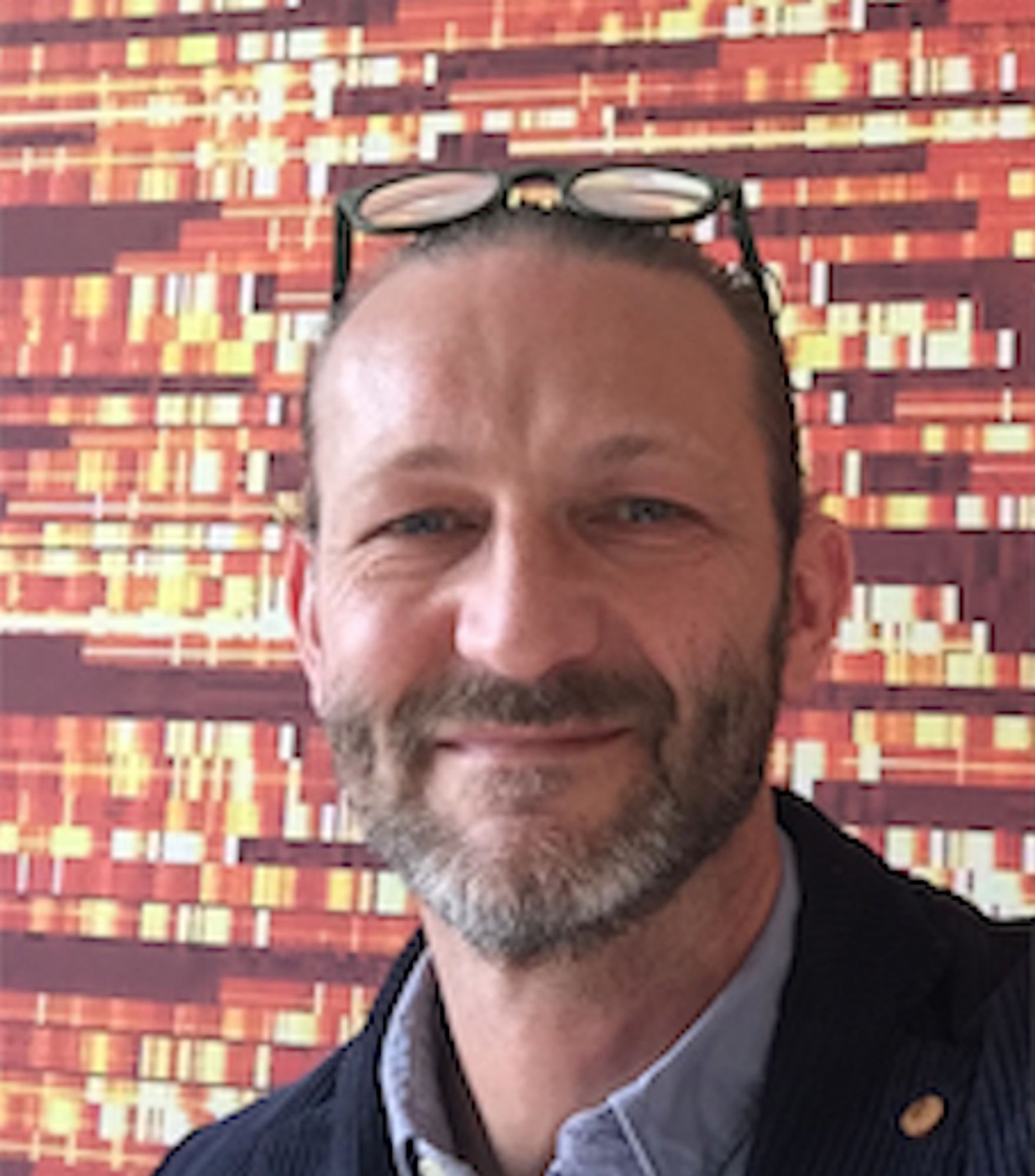 A headshot of a man with dark short hair, a dark beard and a pair of glasses on his head. He is standing in front of a patterned wall of orange, brown and yellow.