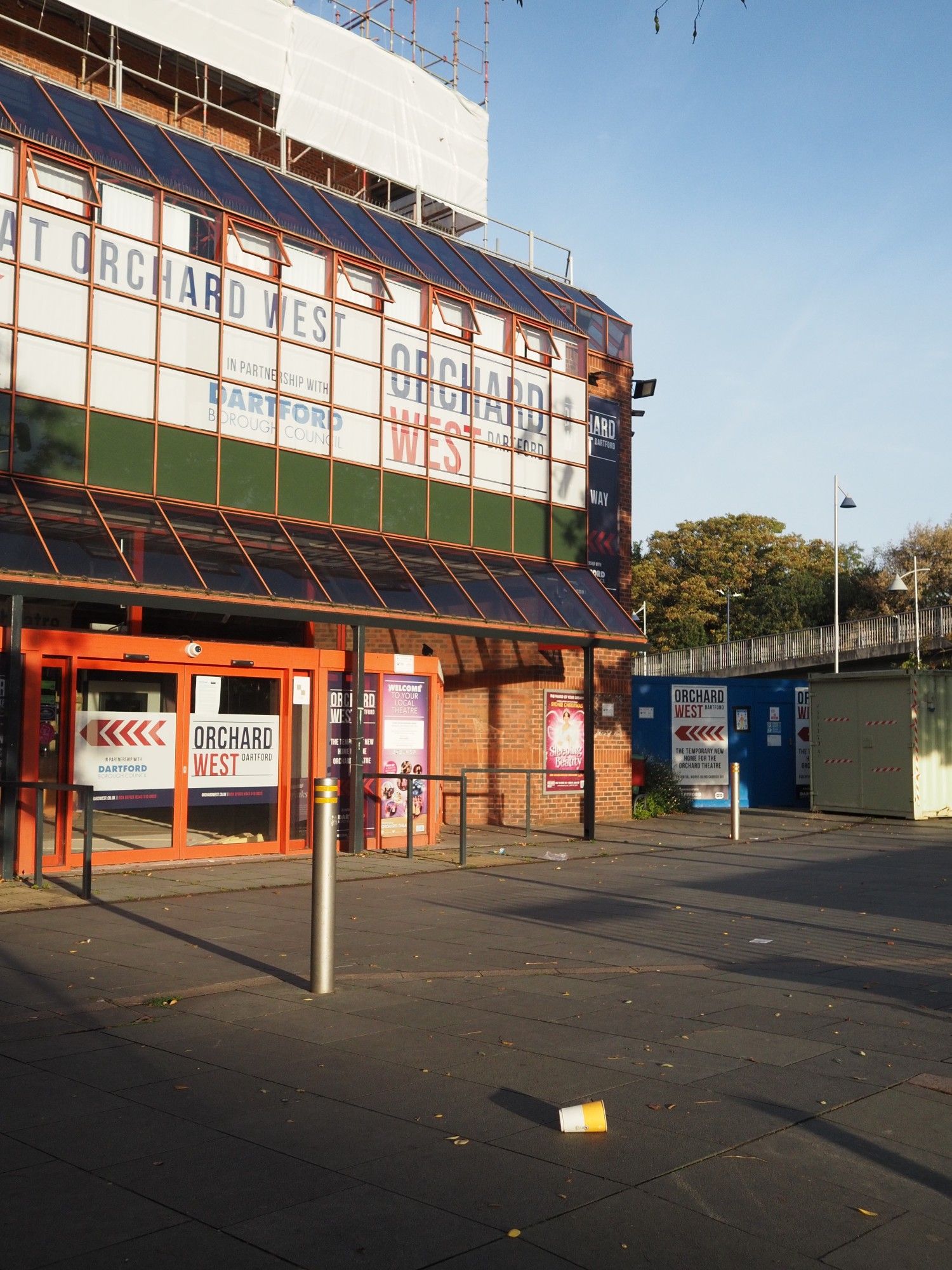 Photograph of a building (a theatre) with a brick and glass frontage. A golden early morning sun lights the building, highlighting the red bricks and red door & window frames. Long shadows of street furniture. On the floor in the centre is a yellow & white takeaway cup.