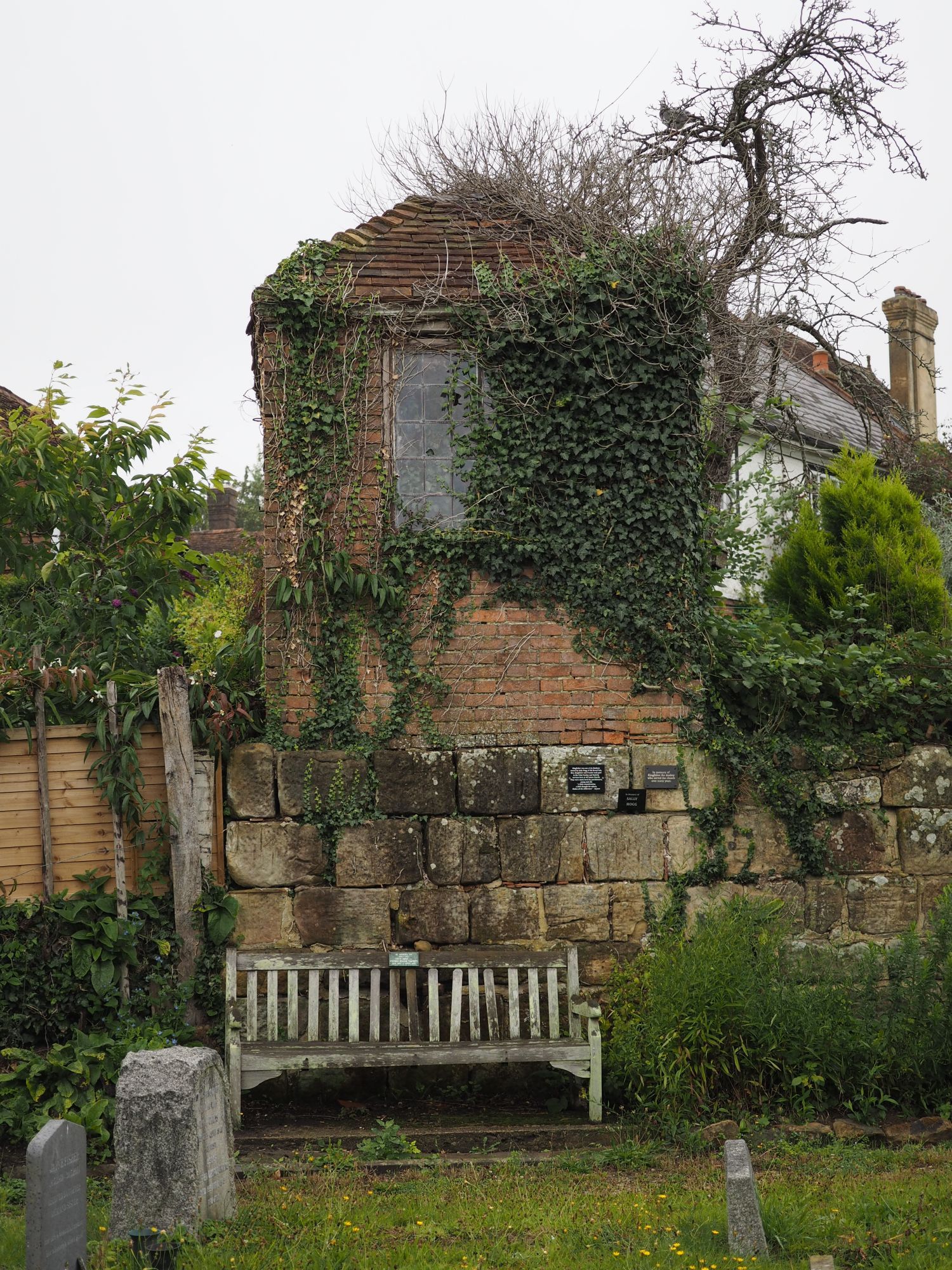 Photograph looking over a few small headstones to a bench facing us. Behind the bench a brick building stands behind a wall. Ivy is creeping around the brick, across an opaque glass window and up onto the roof. A woody shrub towers even higher