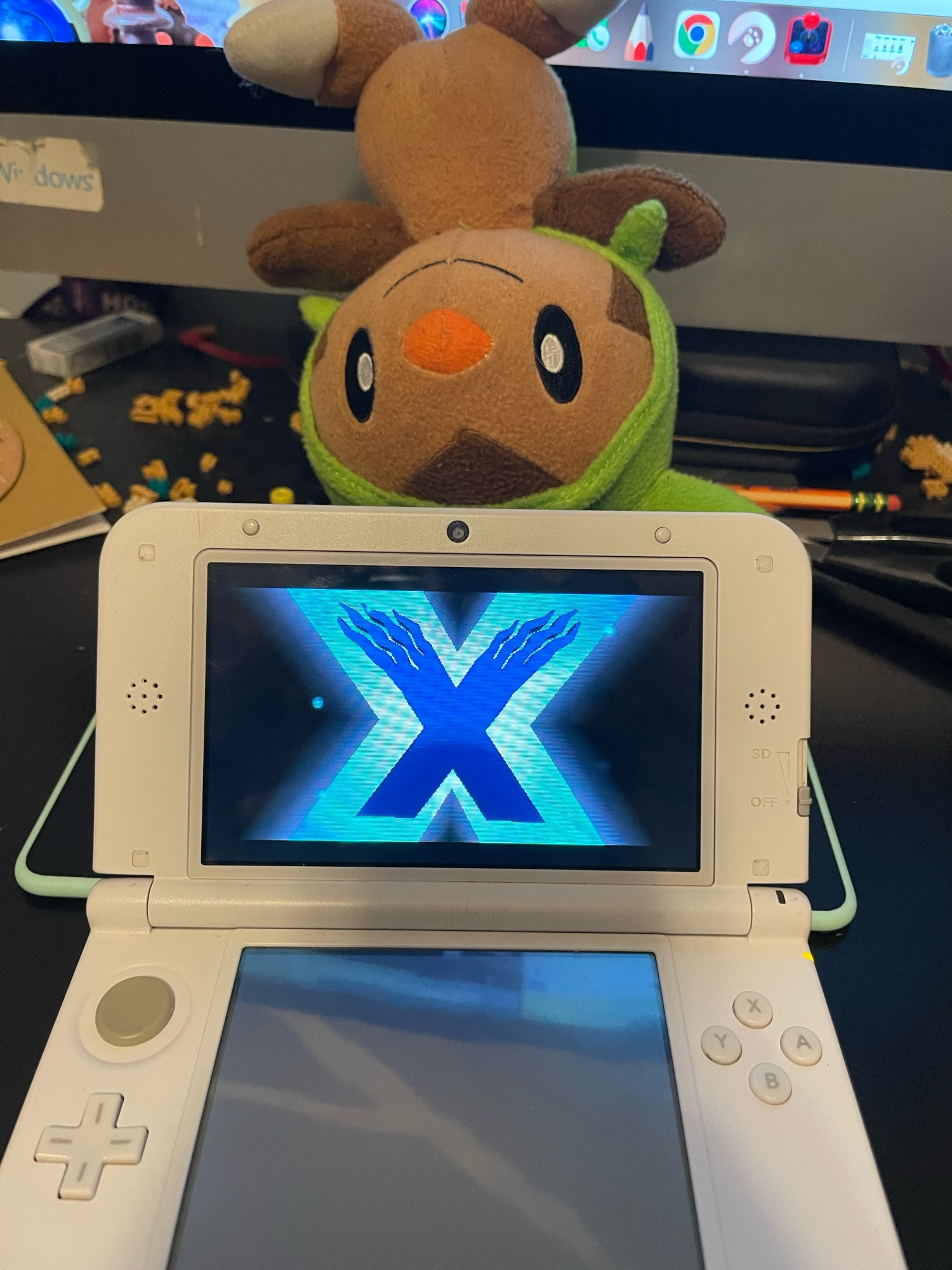 A 3DS with the game pokemon x booted up, with the pokemon chespin as a plushie above