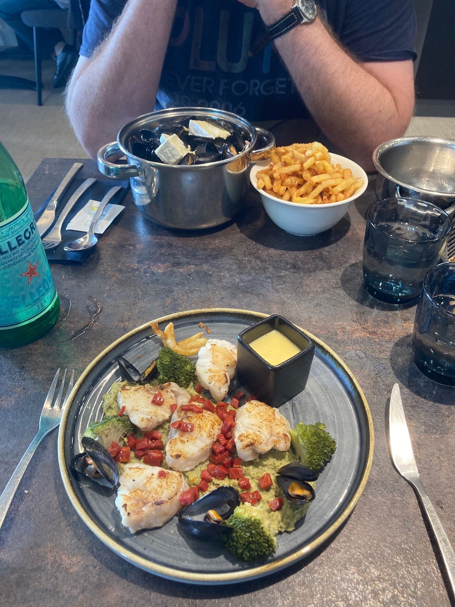 A plate of monkfish on broccoli purée with mussels sits opposite a silver pot of mussels and bowl of frite.