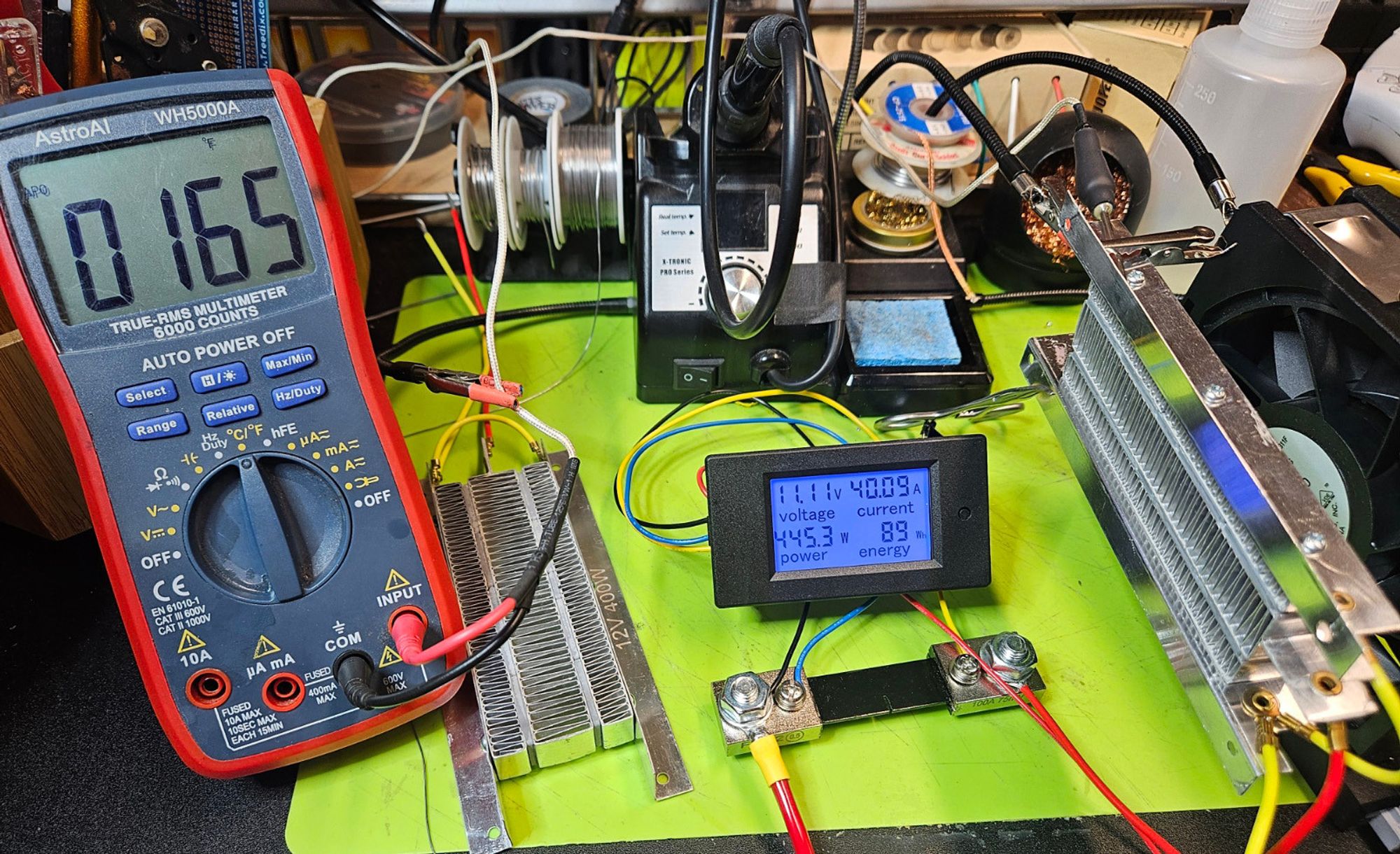 Electronics test bench. A PTC heating element is held in front of a fan. A current shunt displays a power draw of 445 watts (11 volts, 40 amps) and a multimeter reads a temperature of 165 degrees fahrenheit. Another heating element sits on the table unconnected.