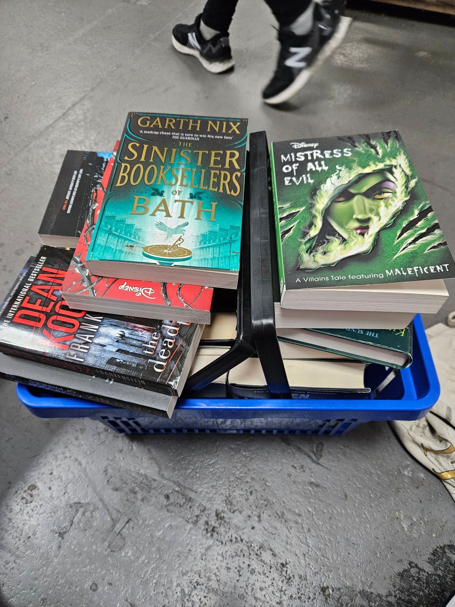 A blue shopping basket, sat on a grey floor. Piled to overflowing with a range of books