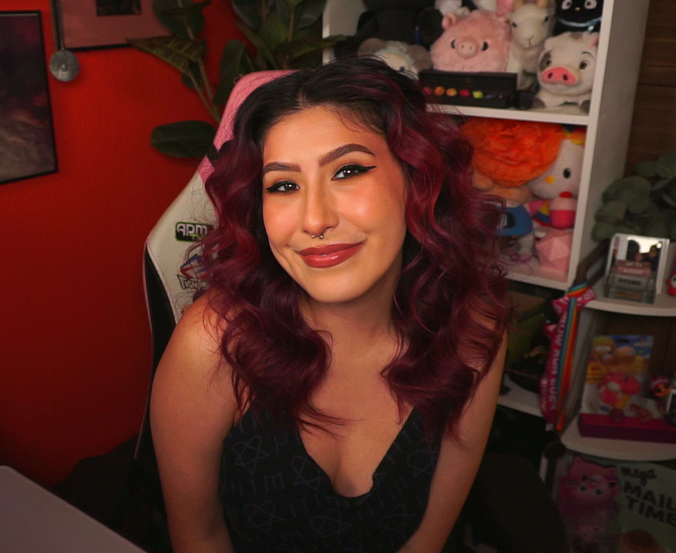 Bailee, a white Latina woman, sitting and posing at her desk. She is wearing a black tank top with purple print on it. Her hair is deep red and curled. 