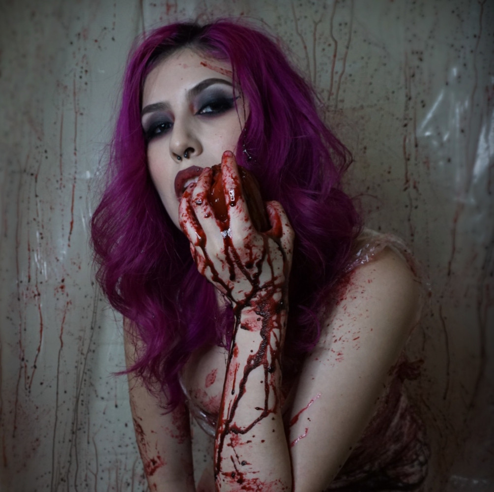 A Halloween photoshoot of Bailee where she is appearing to eat a heart. Blood is splattered all over the walls and dripping down her hands. She is wearing a clear, form fitting dress. Her hair is a deep magenta and curled.