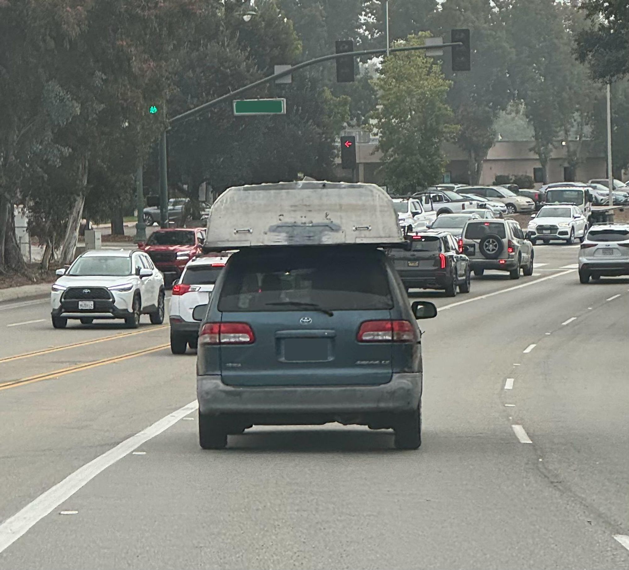 A blue toyota minivan drives down a road with a large upside-down row boat riding on top of the van’s roof. No methods of securing the boat are visible, and it is riding directly on top of the van’s roof.

Other cars are visible on the road beyond it.