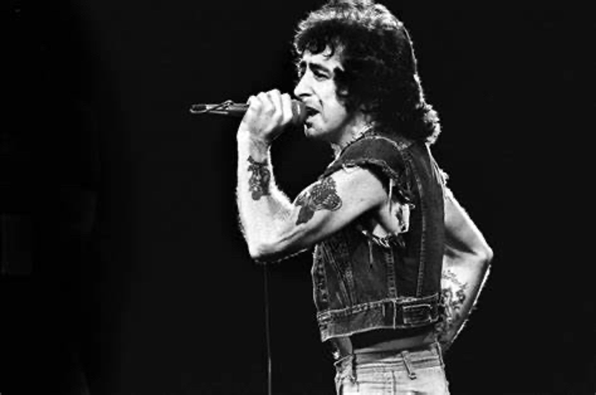 Black and white photo of original AC/DC singer Bonn Scott singing. He’s wearing a cut off denim jacket and singing into a microphone.