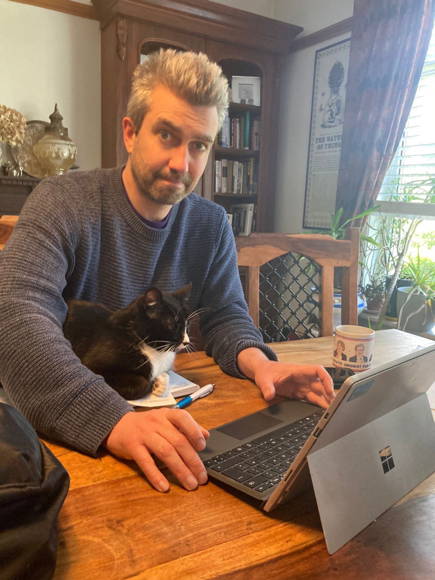 Danny sitting at a table in front of a laptop with a black and white cat under his arm.