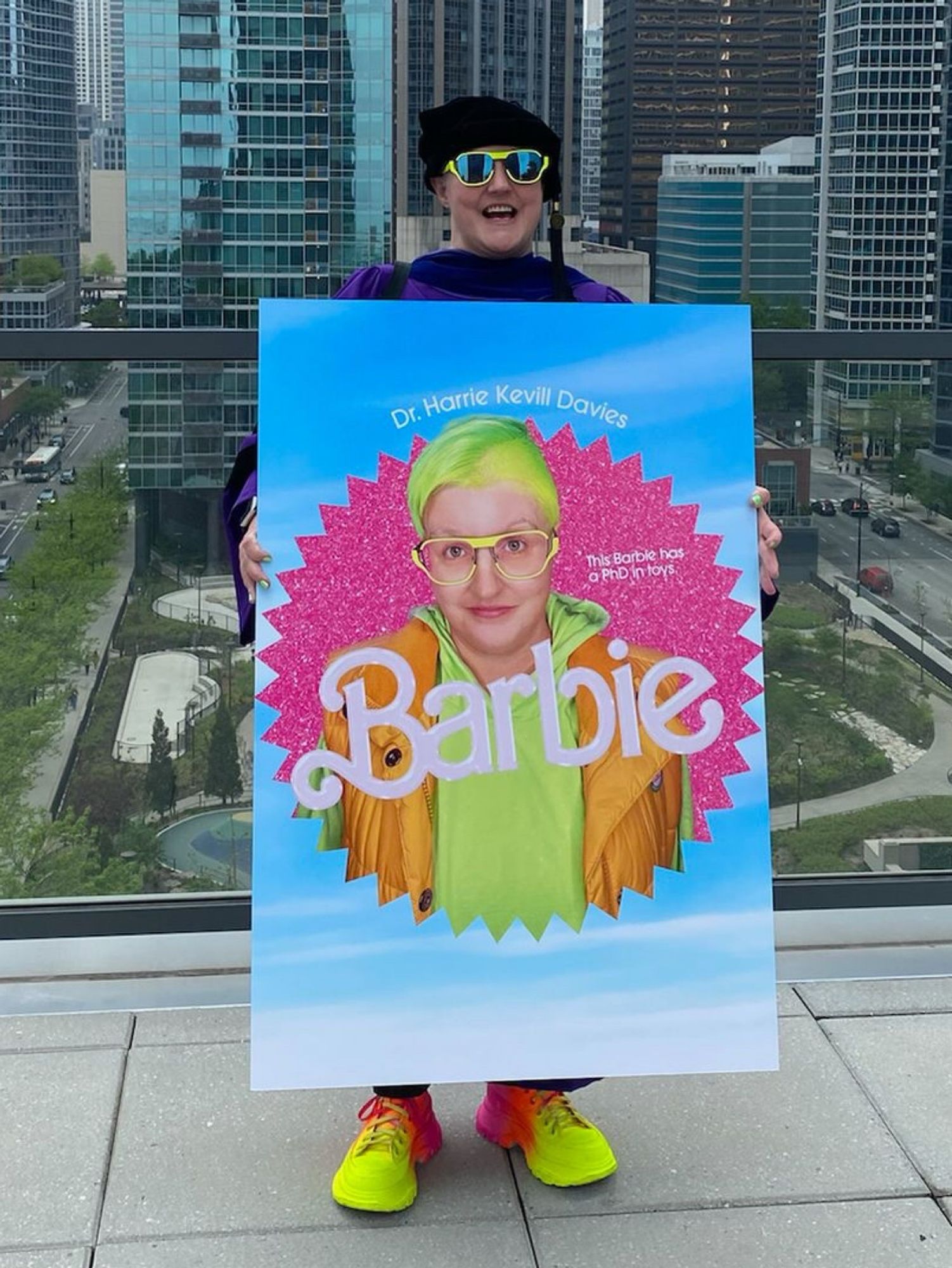 A picture of my friend Harrie at their hooding ceremony in 2023. They’re standing in their doctoral regalia on their building’s patio, holding the custom Barbie movie poster that Mattel made especially for them.