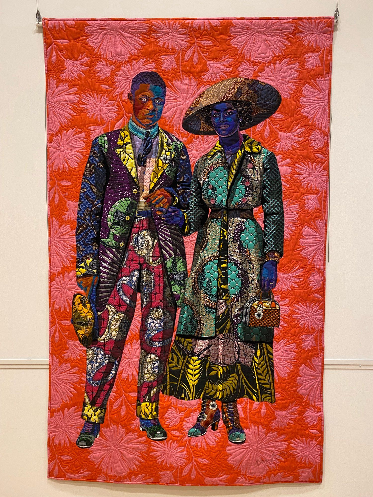 The whole work, depicting a man and woman holding hands. The figures are depicted using layered, brightly colored, beautifully patterned fabrics against a pink and orange-red patterned background. The colors and joy expressed through them reflect the joyful subject matter: a vibrant reimagining of a photograph of 1930s Black Americans commemorating the event of their marriage with a photograph.