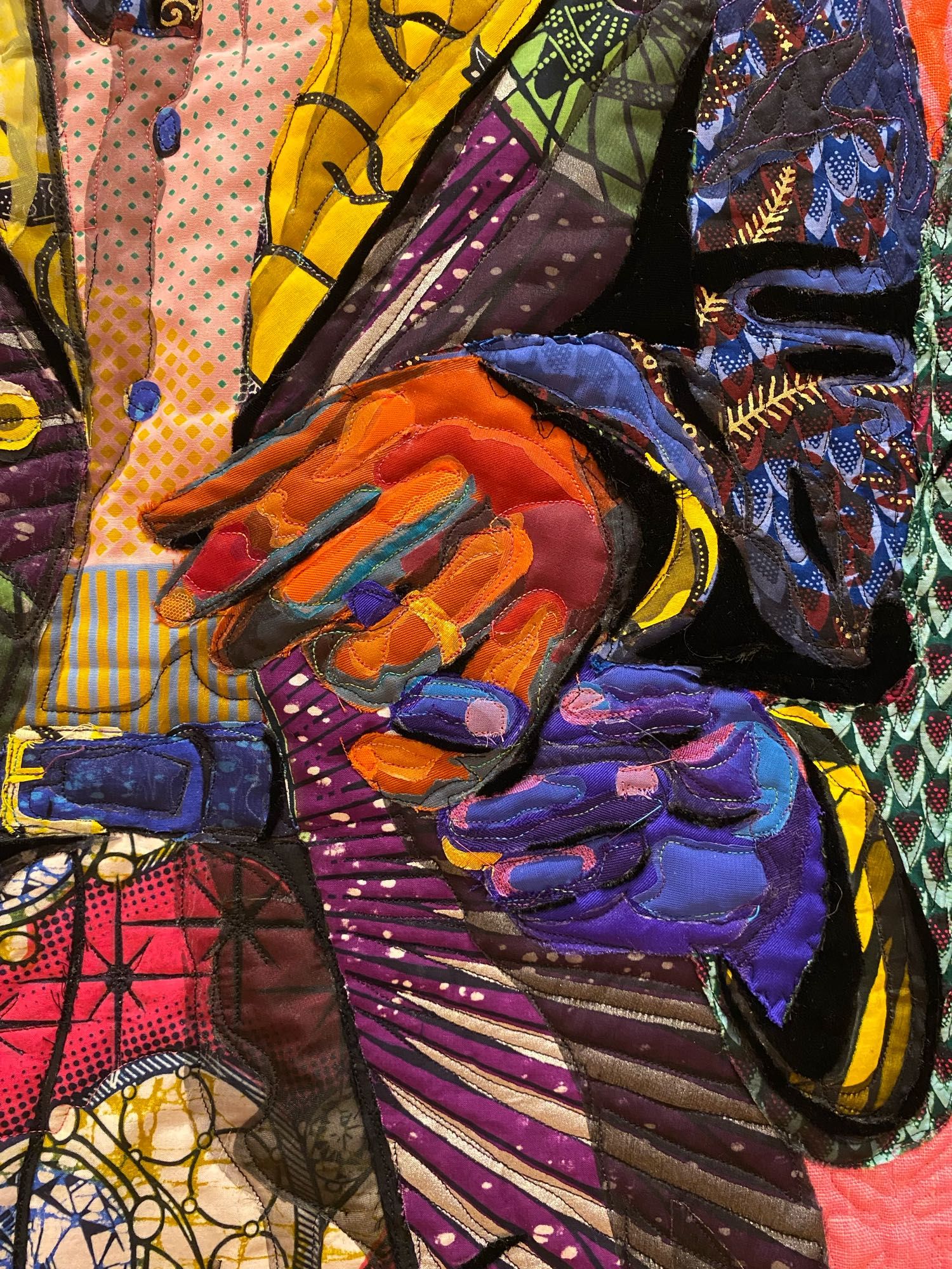 Detail of entwined hands: his, left, wearing a wedding ring; hers, right, lovingly grasping his little finger. The hands and figures’ clothing are depicted using layered, brightly colored, beautifully patterned fabrics.