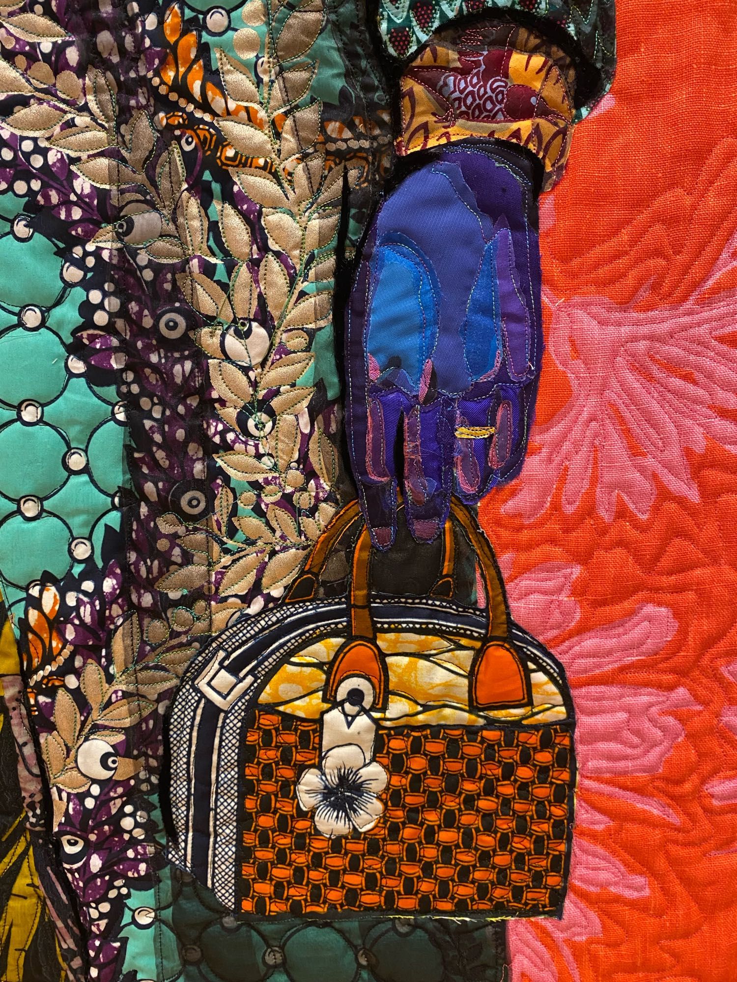 Detail of the woman’s left hand, wearing a wedding ring and holding a handbag, depicted using layered, brightly colored, beautifully patterned fabrics against a vibrant pink and orange-red background fabric.