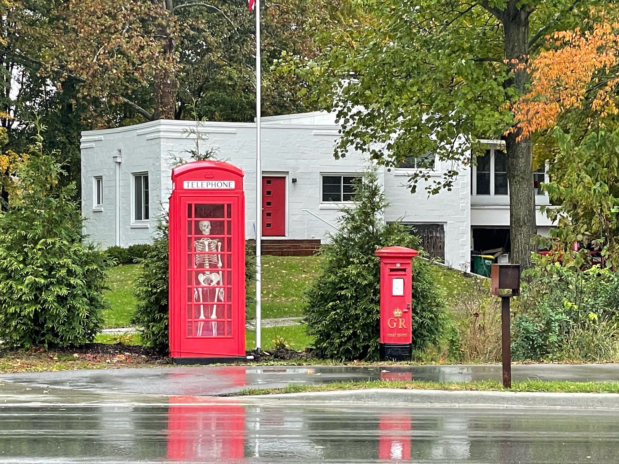 A skeleton standing inside a phone box that is next to a letter box. A white house with a red door is in the background 