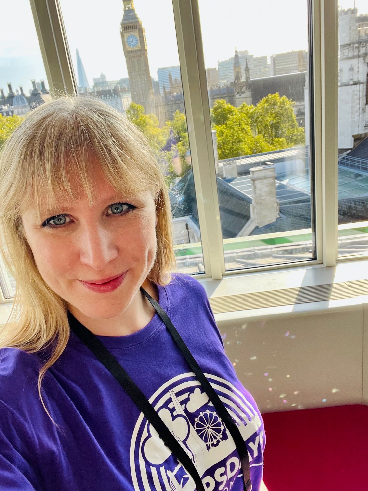 Hannah is wearing a purple DevOpsDays T-shirt and is standing in front of an amazing view of Big Ben