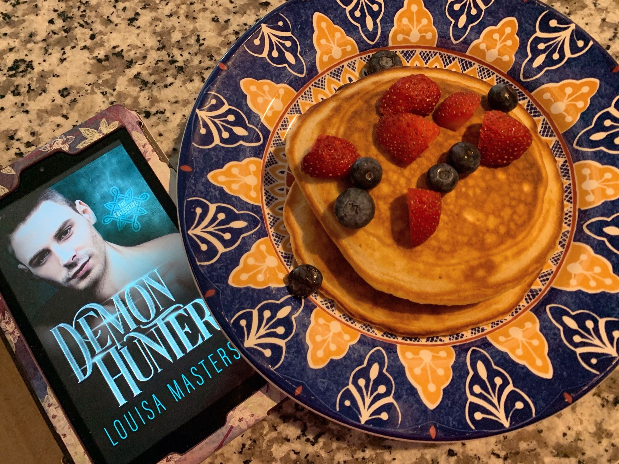 A kindle showing the cover of Demon Hunter on the screen next to a plate of pancakes