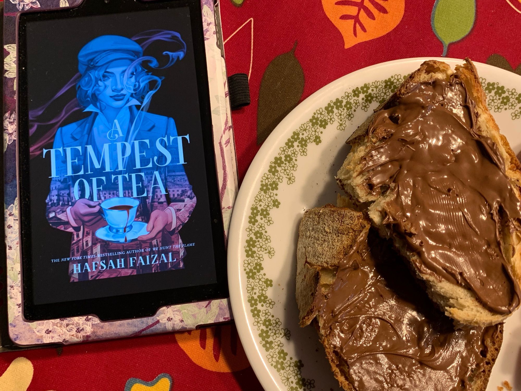 A kindle showing the cover of A Tempest of Tea next to a plate of toast with Nutella