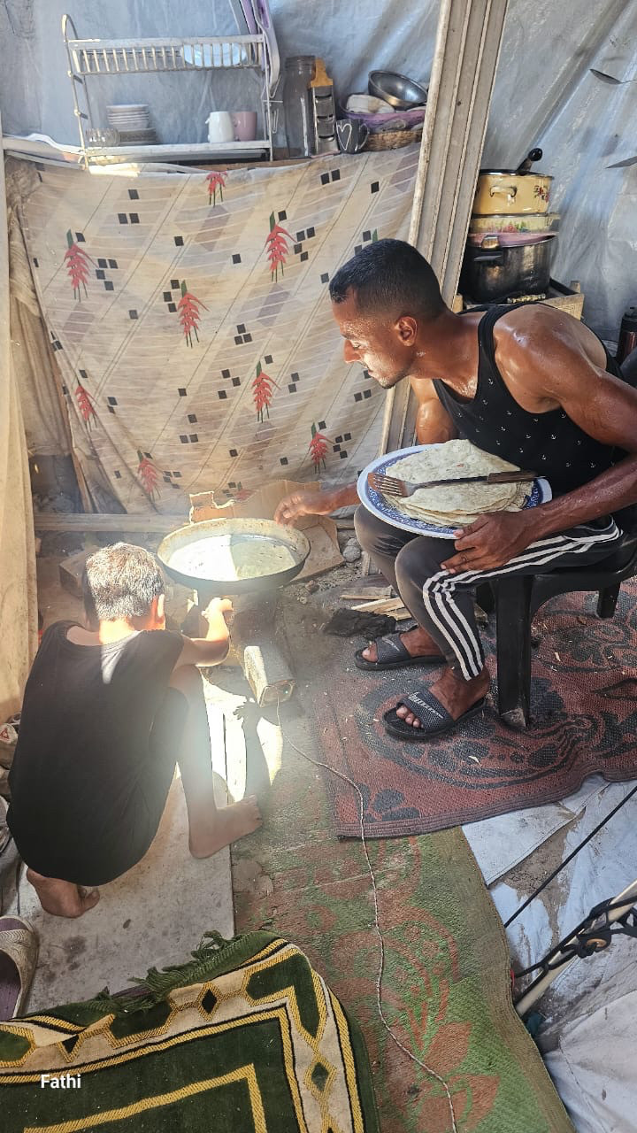 Fathi sits on a chair hunched over a camping stove with a plate of shrak bread in his lap. He is visibly sweating from the sun’s heat inside tent and the heat of the stove. Fathi’s son Yamen crouches with his back to us as he monitors the flame in the stove.