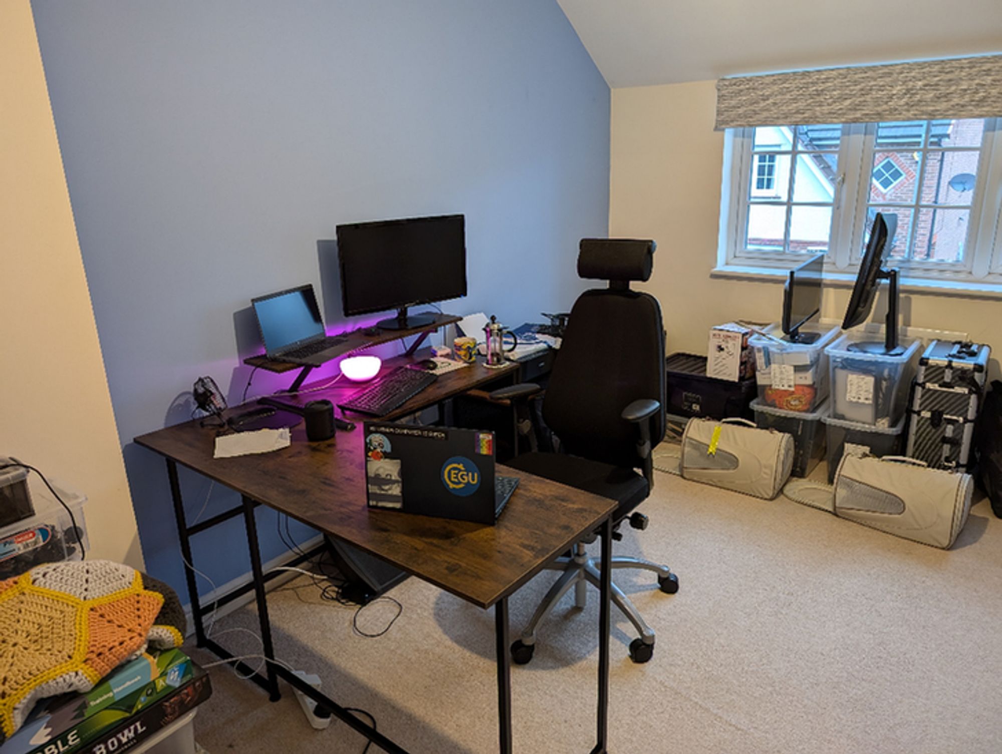 A messy home office. A corner desk and office chair, two laptops, and a coffee. Surrounded by boxes.