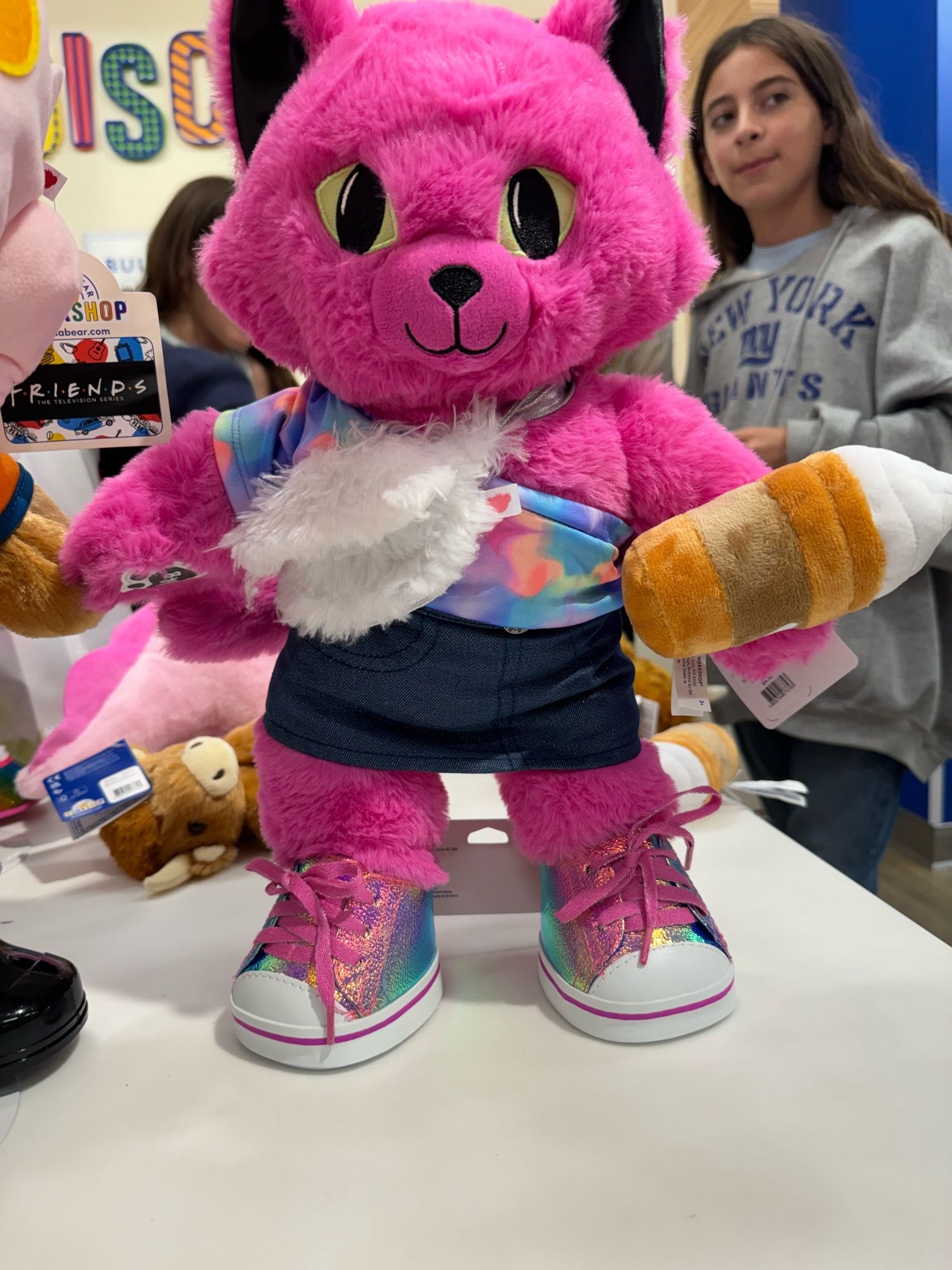 alarmingly pink stuffed cat, wearing an off the shoulder neon print top, a denim skirt, and metallic ombré shoes, carrying a white fluffy fanny pack and a stuffed starbucks beverage