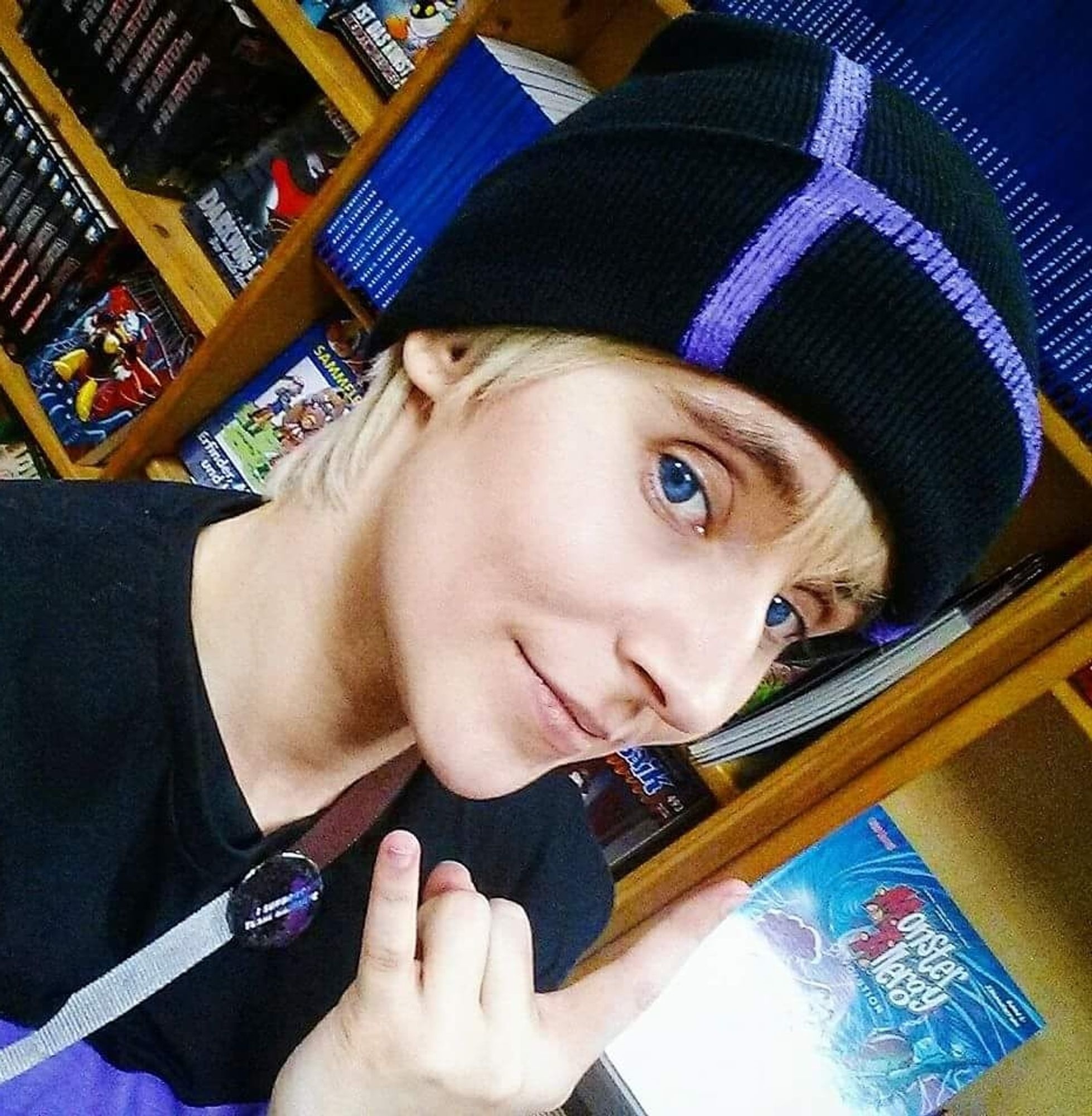 A whitw person in their 20s with a black beanie, black shirt and blue eyes. They wears the blonde wig beneath the beanie