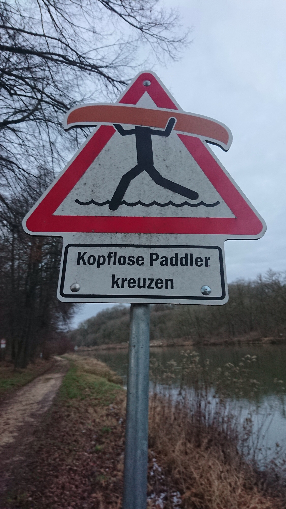 „Kopflose Paddler kreuzen“ ist der Text auf einen weißen Schild unterhalb eines rot-weißen dreieckigen Warnschild, mit einem skizzierten Mensch mit dem Kopf unter einem Boot, das sogar über das Dreieck des Warnschilds rechts und links etwas hinausragt.