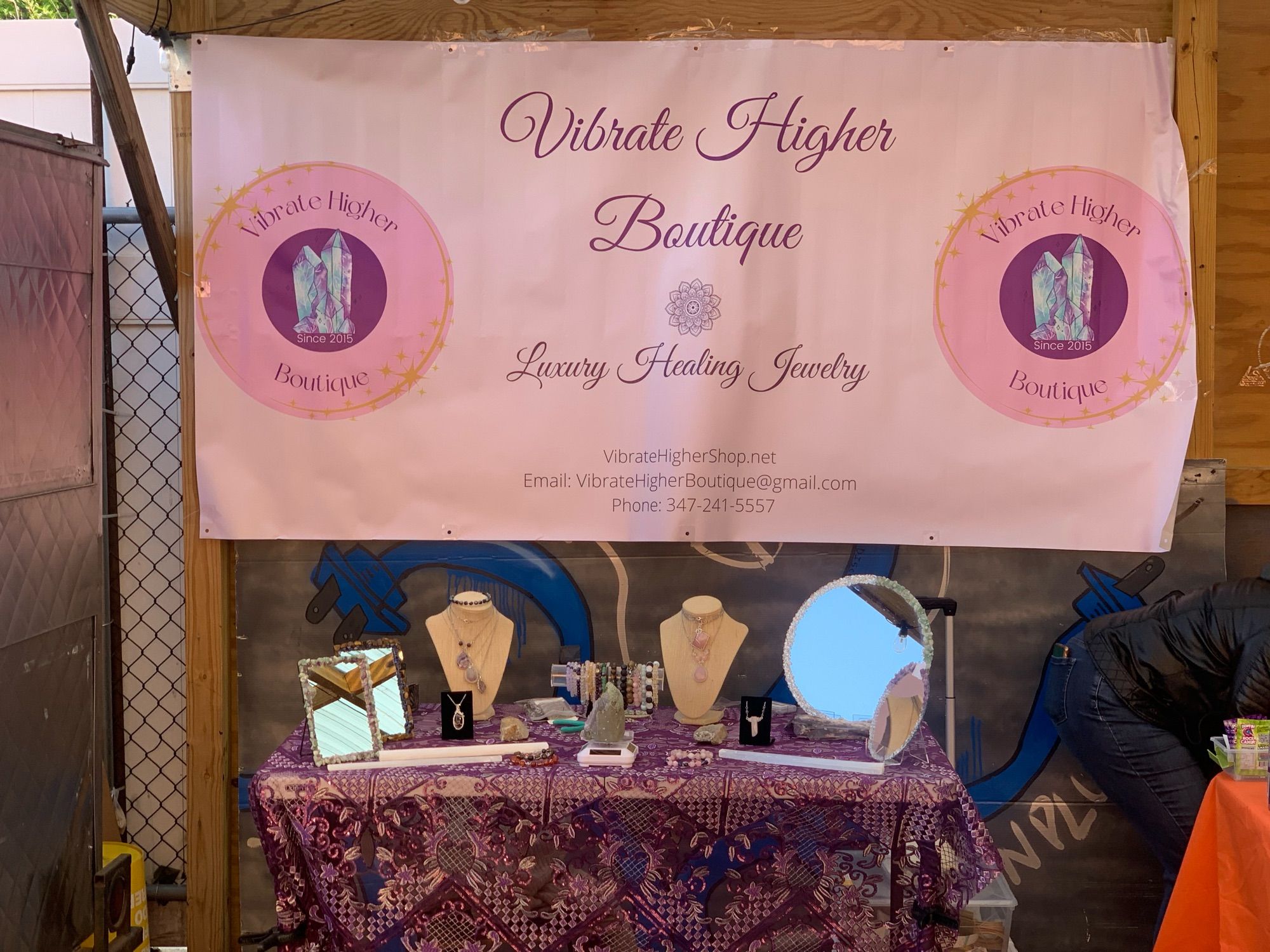 Photo of our tables set up when we do pop up vending. Our table is purple lace with tones of crystal goodies on it! 

Our very pink banner is hanging above, behind the table that has our logo and our name which is “Vibrate Higher Boutique” our slogan is “Luxury Healing Jewelry” because good quality items should be available to everyone