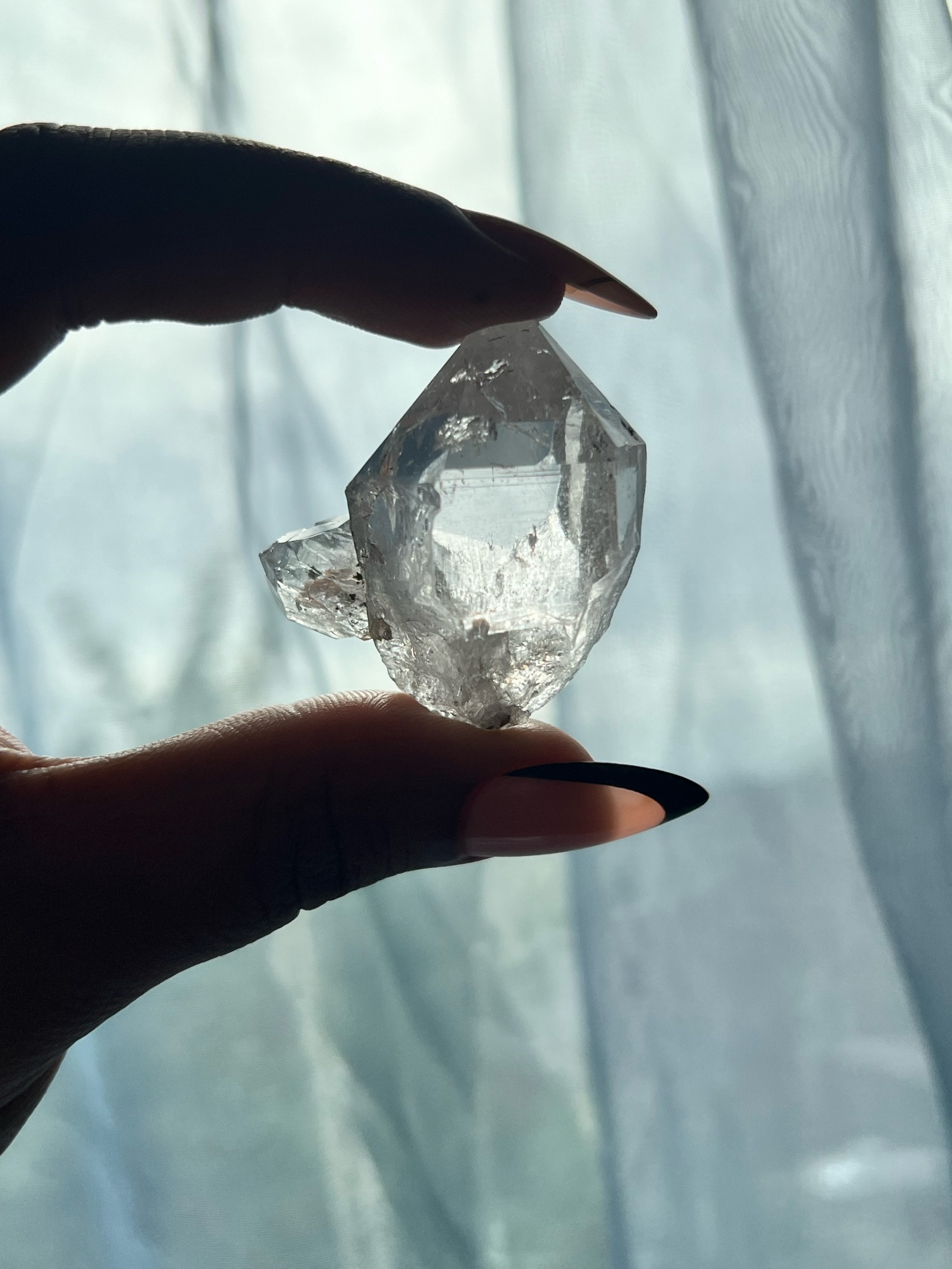 A clear Quartz crystal known as Herkimer Diamond being held up to the light between two fingers