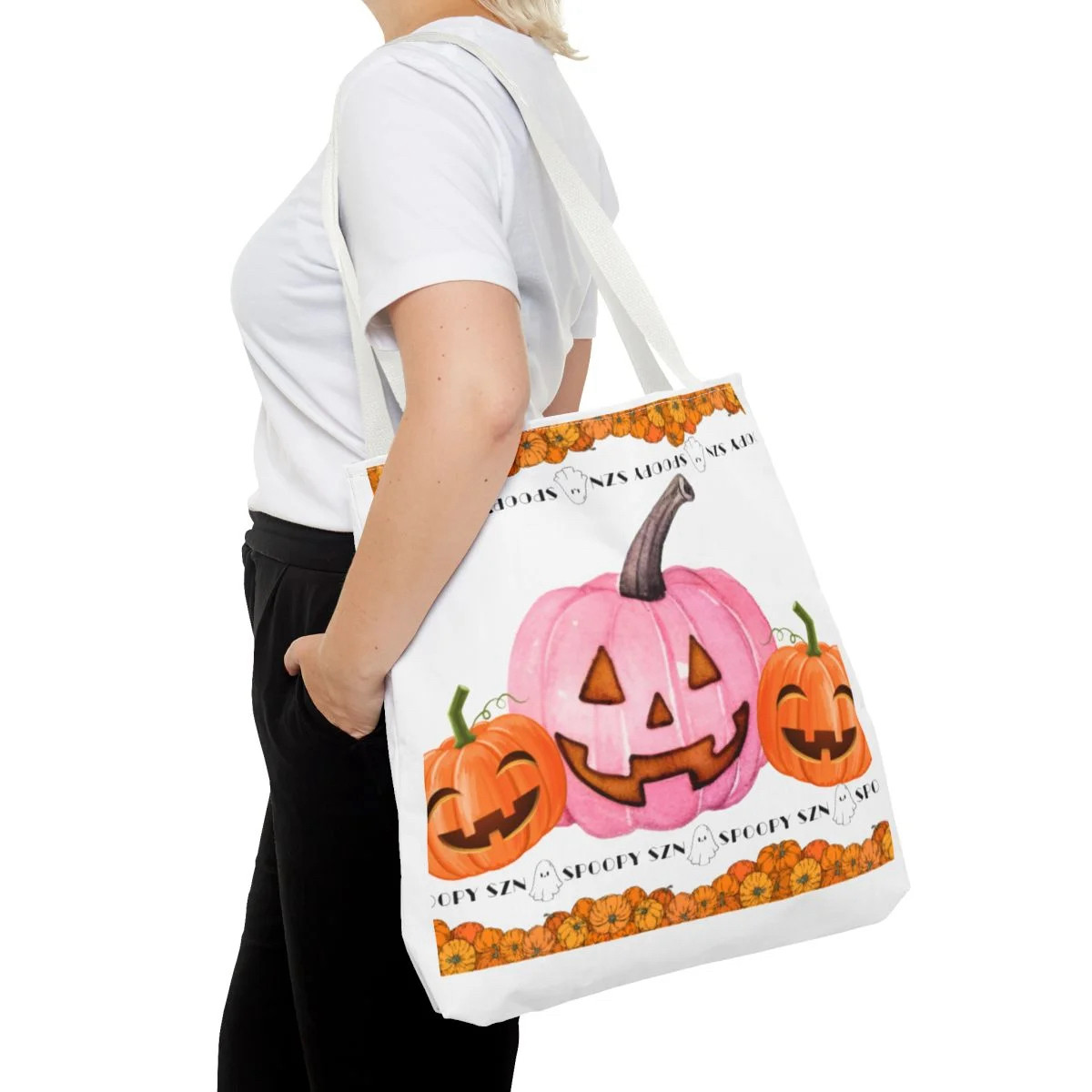 A person shown with the tote bag on their back in the largest size. A white tote bag with pumpkins displayed & a banner that says “Spoopy szn” across the top and bottom