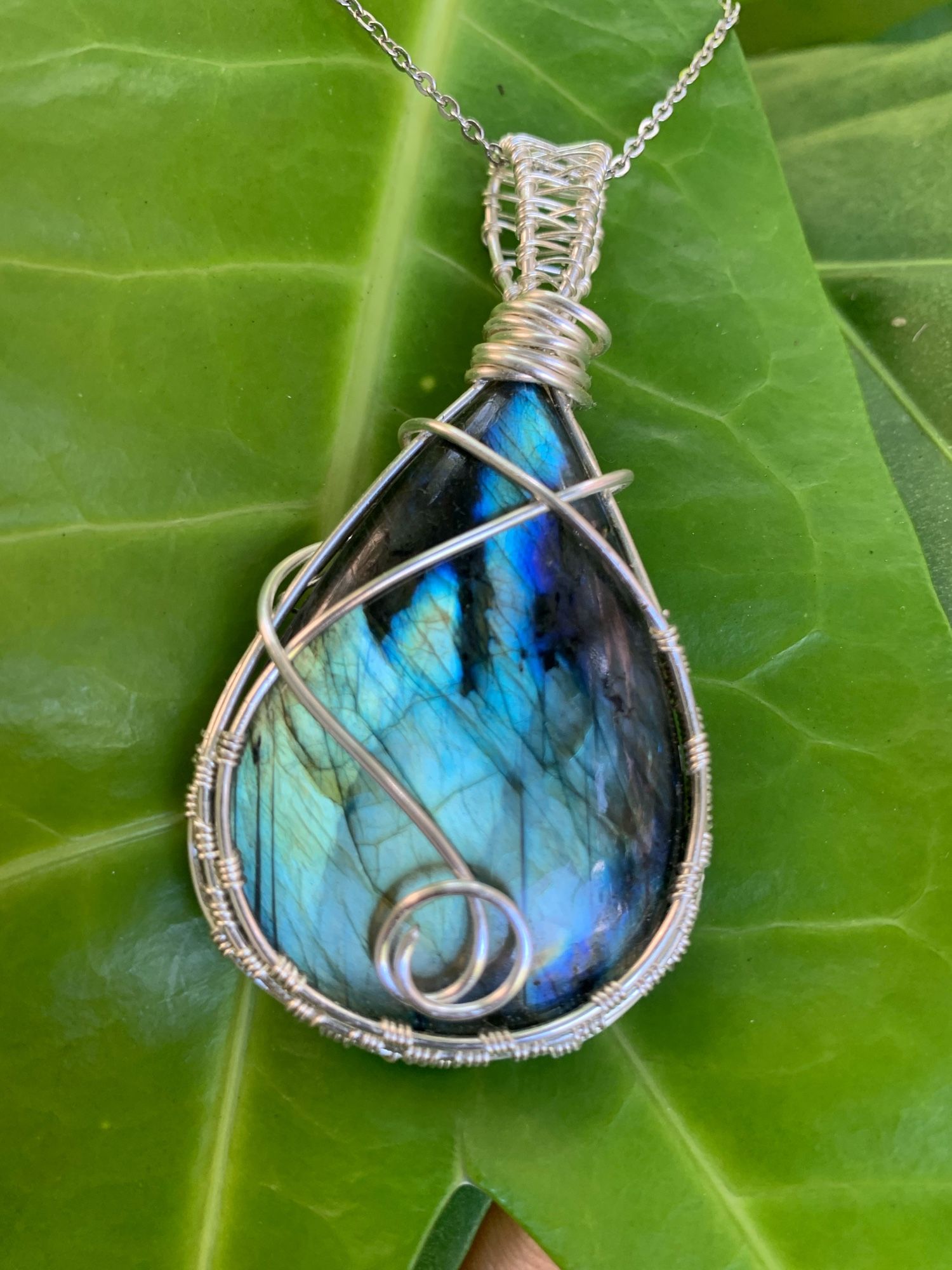 A vibrant blue XXL labradorite with a flash that is shaped like a flame. Wrapped in silver wire and displayed on a large leaf