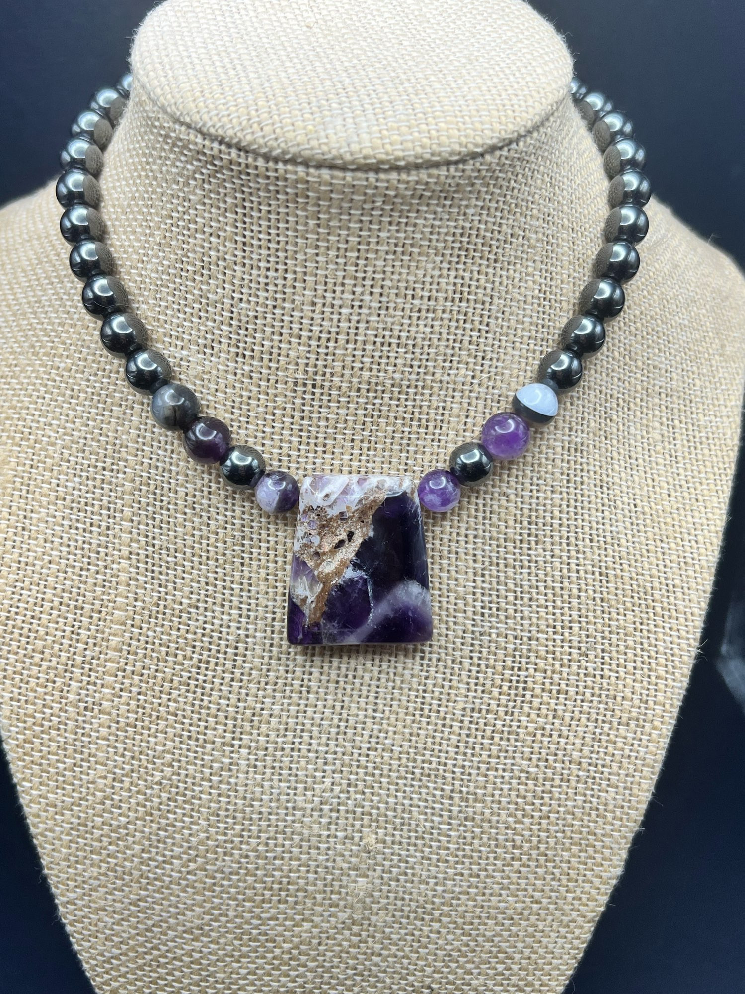 A necklace displayed with silver dale grey hematite beads, opaque black agate beads and light purple amethyst beads with a squareish Chevron amethyst in the center that has white banding and shows some matrix