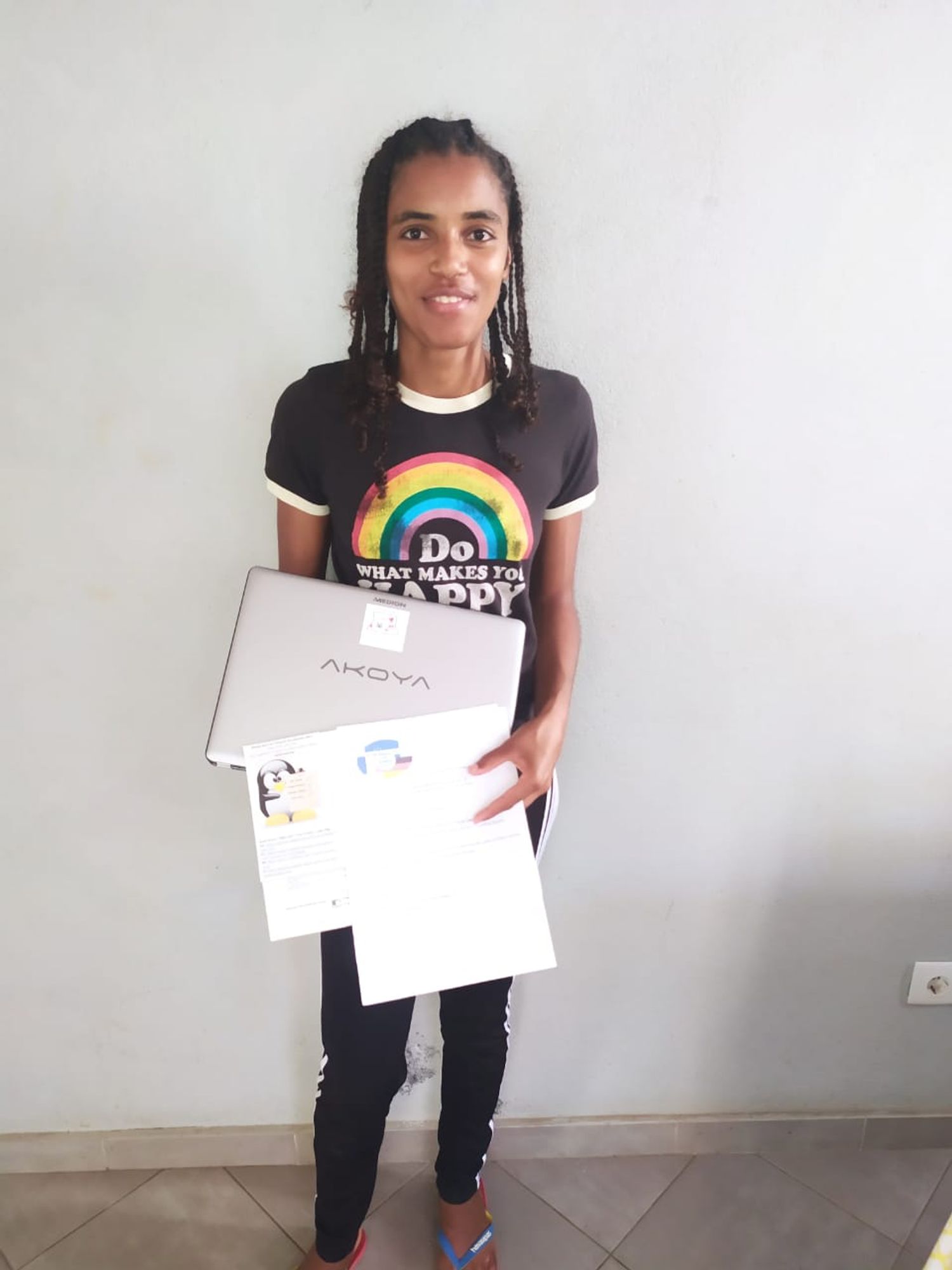 Das Foto zeigt eine junge Studentin vor einer weißen Wand. Sie trägt ein schwarzes T-Shirt mit dem Aufdruck Do what makes you happy und einem Regenbogen darüber. In der Hand hält sie einen Laptop von Labdoo mit ausgedruckten Anleitungen. Sie lächelt in die Kamera.