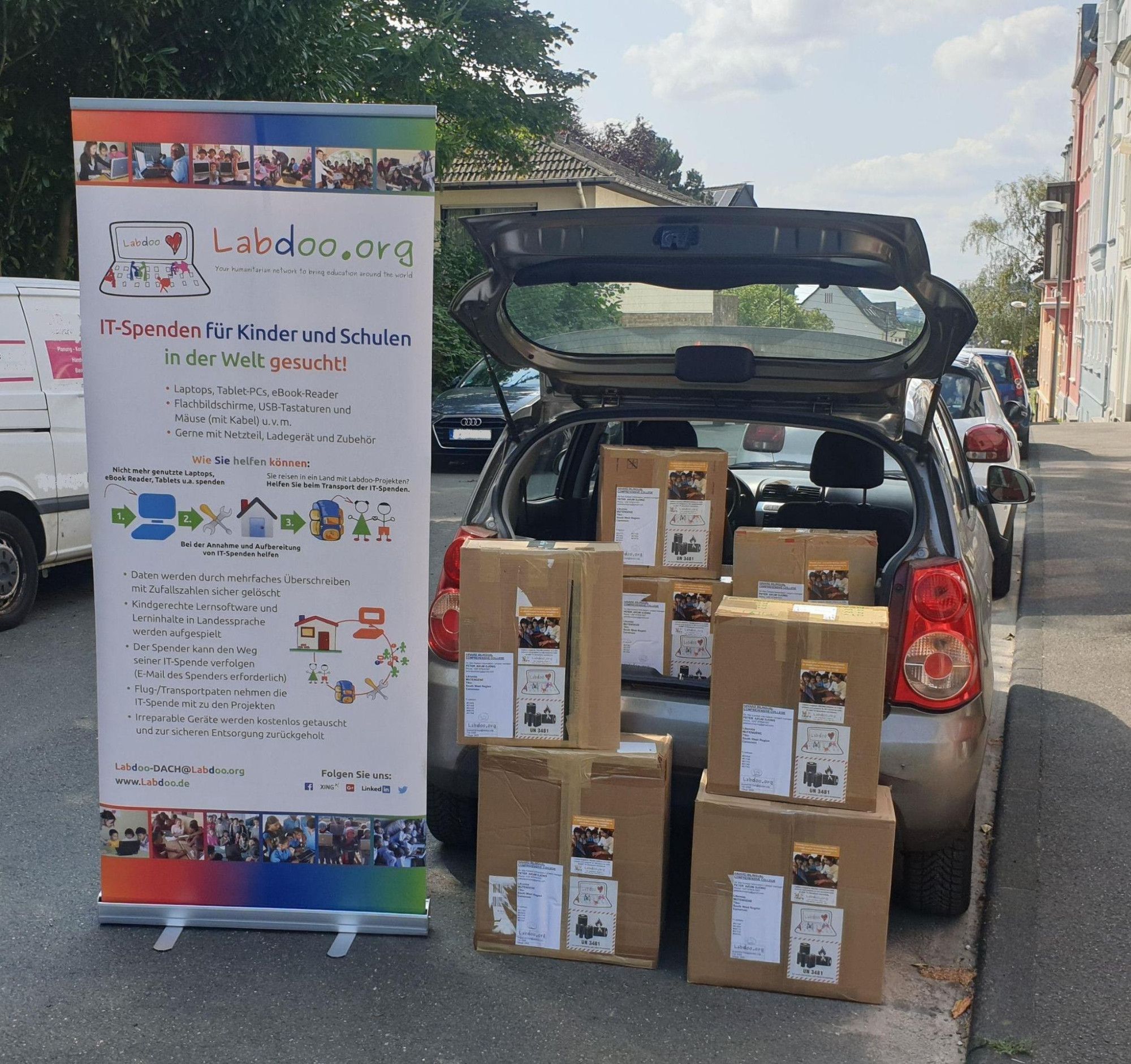 In einer Straße in Wuppertal steht ein Auto mit aufgeklappter Heckklappe- Davor und auch schon eingeladen steht ein kleiner Berg von ca. 8 Paketen und ein Rollup von Labdoo.