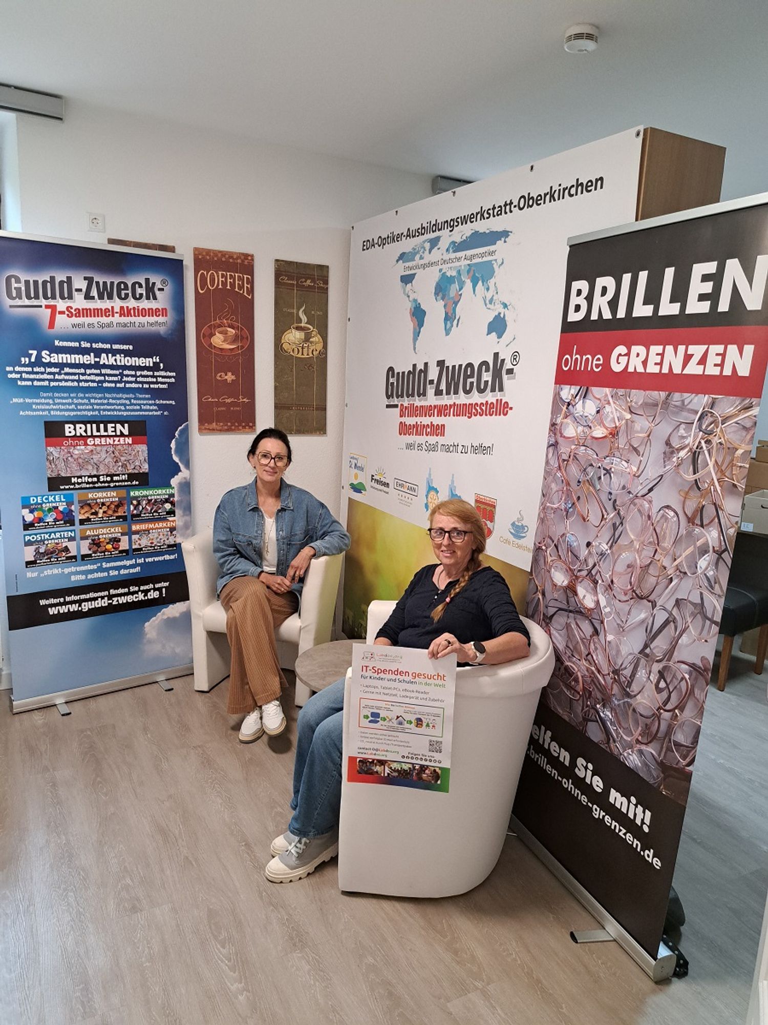 In einer Raumecke in einem Café sitzen in hellen Sesseln 2 Frauen, Micheala con Brillen ohne Grenzen und Hanny von Labdoo. Im Hintergrund hängen vor einer weißen Wand Plakate der Projekte. Beide Frauen schauen in die Kamera und lächeln. Hanny hält noch ein Poster von Labdoo in der Hand.