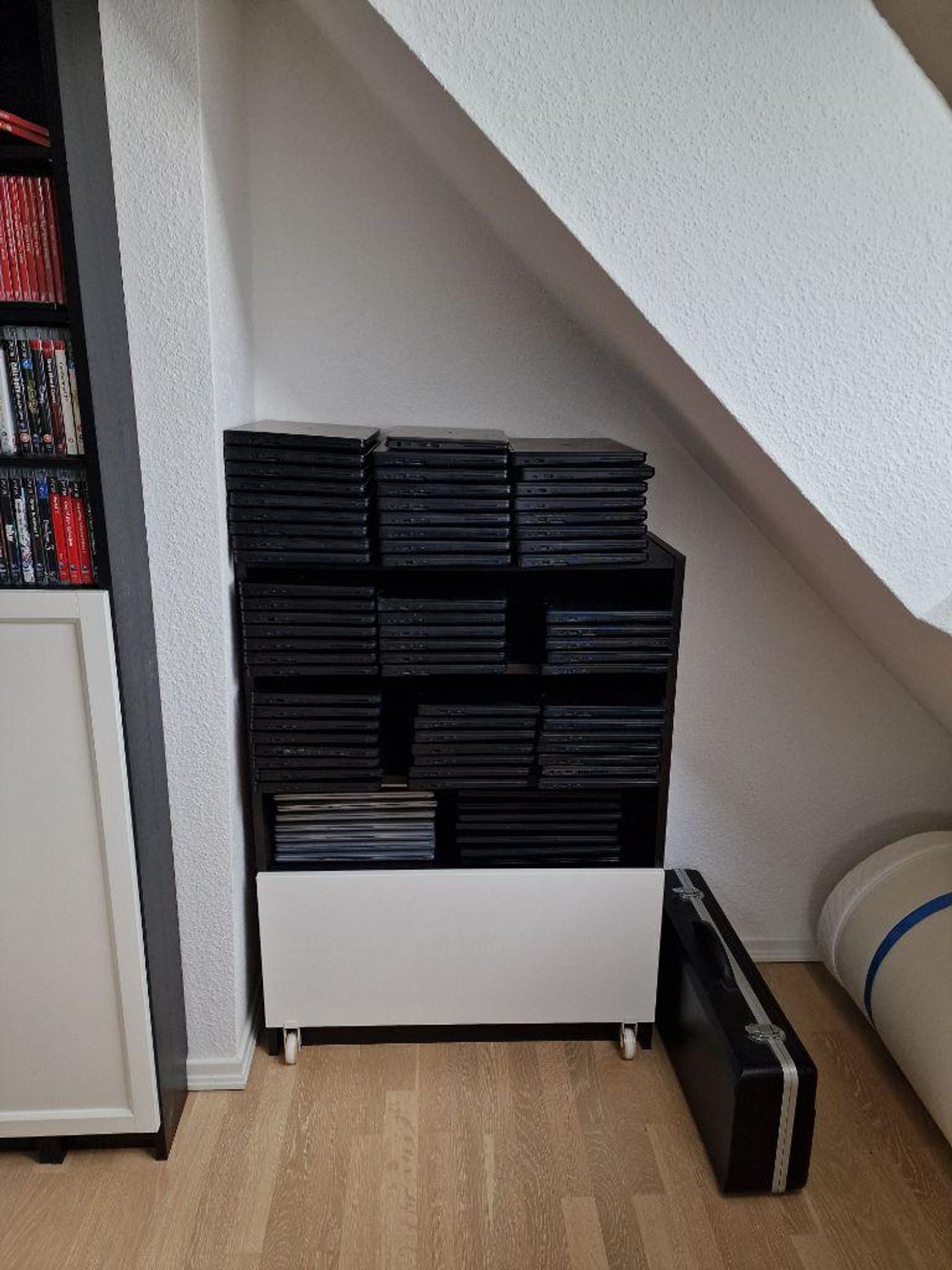 In einer weißen Regalwand unter einer Dachschrägen liegen mehrere Stapel Laptops aufeinander. Darunter ein weißes Sideboard, rechts davon ein Koffer.