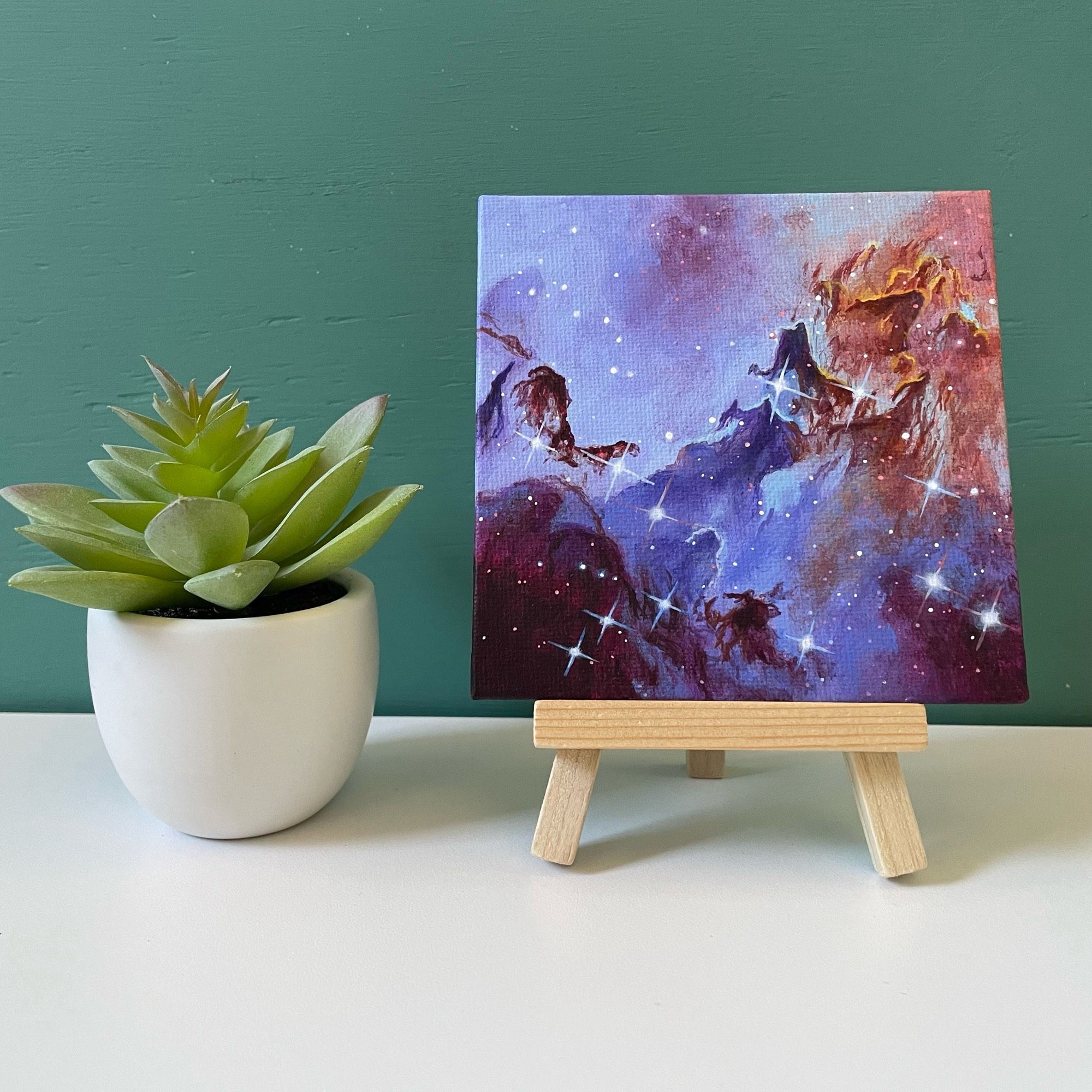 Mini painting of pink and purple nebula on easel next to fake plant