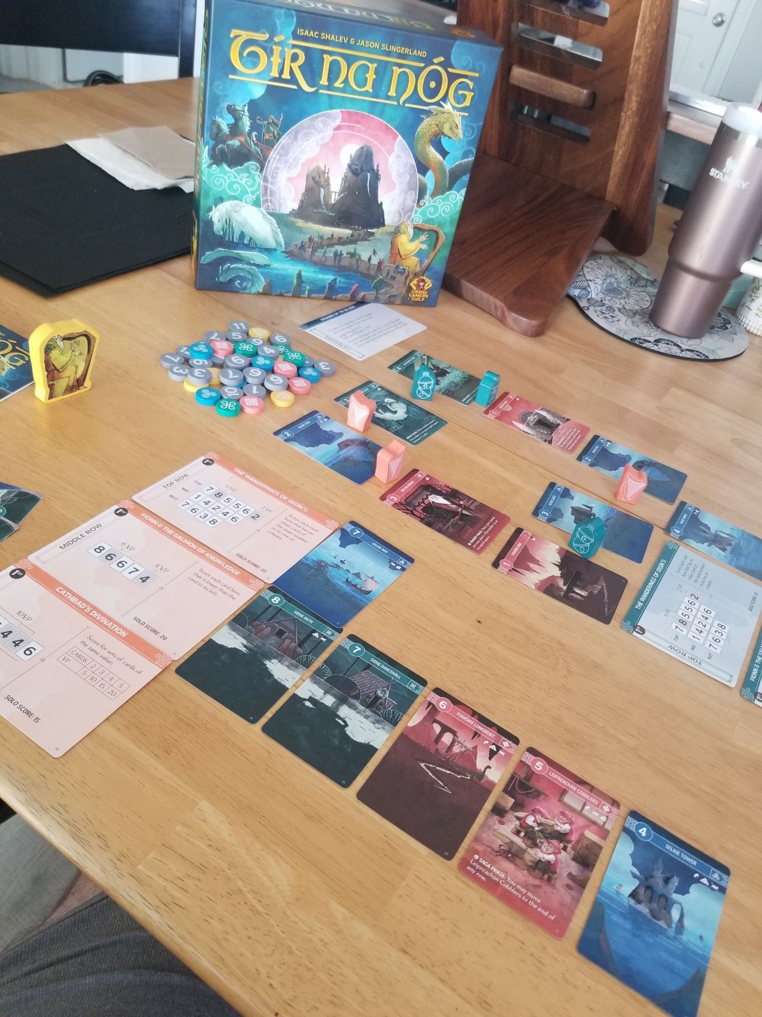 A game of Tír na nÓg in progress. A tableau of colorful cards depicting scenes from Irish mythology. Big chonky wooden bits. Gorgeous box art featuring the road to the Celtic Otherworld. Jenn's rose gold Stanley cup is on the table too.