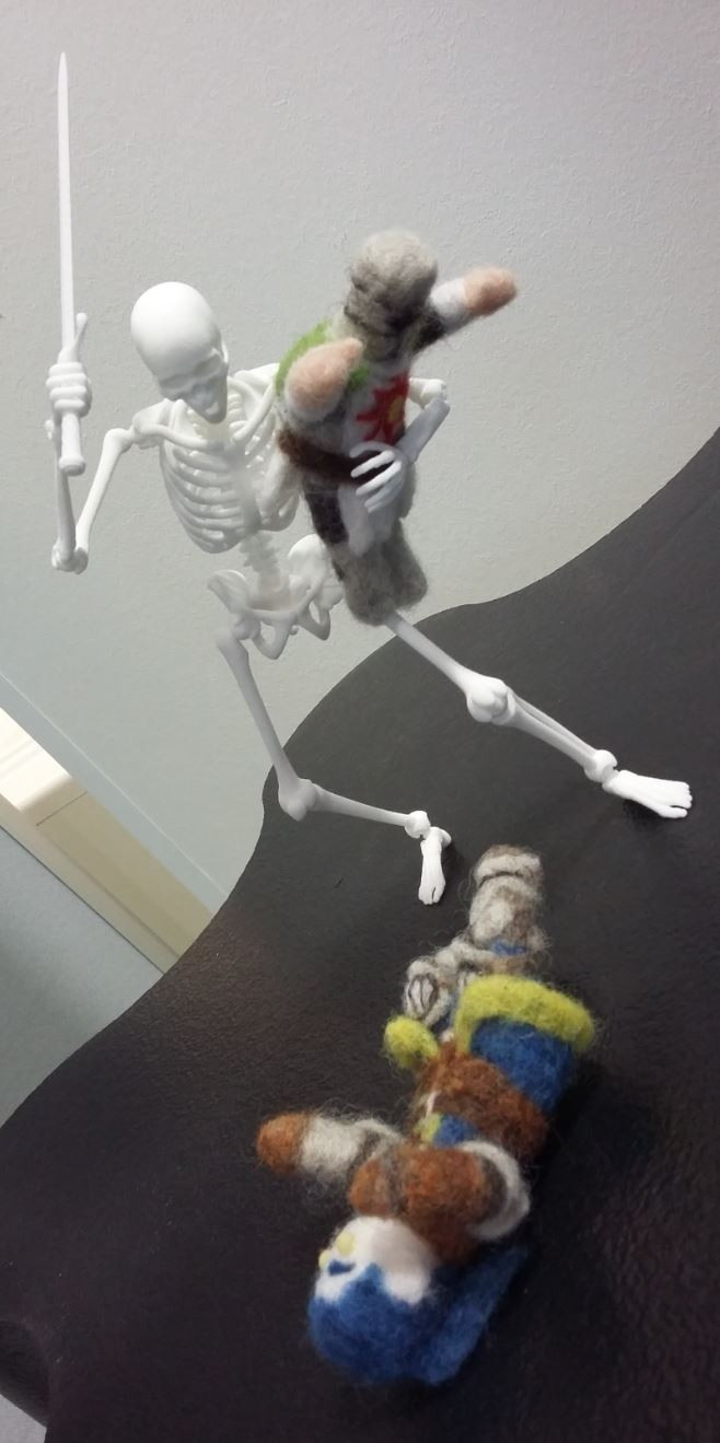 A photo of two needle-felted characters, one my own character, Zesil, dressed up in the elite knight armour from Dark Souls, and the other is of Solaire of Astora, a friendly sun-worshipping knight also from Dark Souls.
The two are fighting against a giant, 15 centimetre tall 3D printed skeleton! Or, rather, Zesil rolls on the ground, gasping as the skeleton raises its sword high up while holding the tiny Solaire in his arm like a body pillow.
As fierce as the skene may seem, the three figurines are actually good friends! I guess they just like acting for the camera...