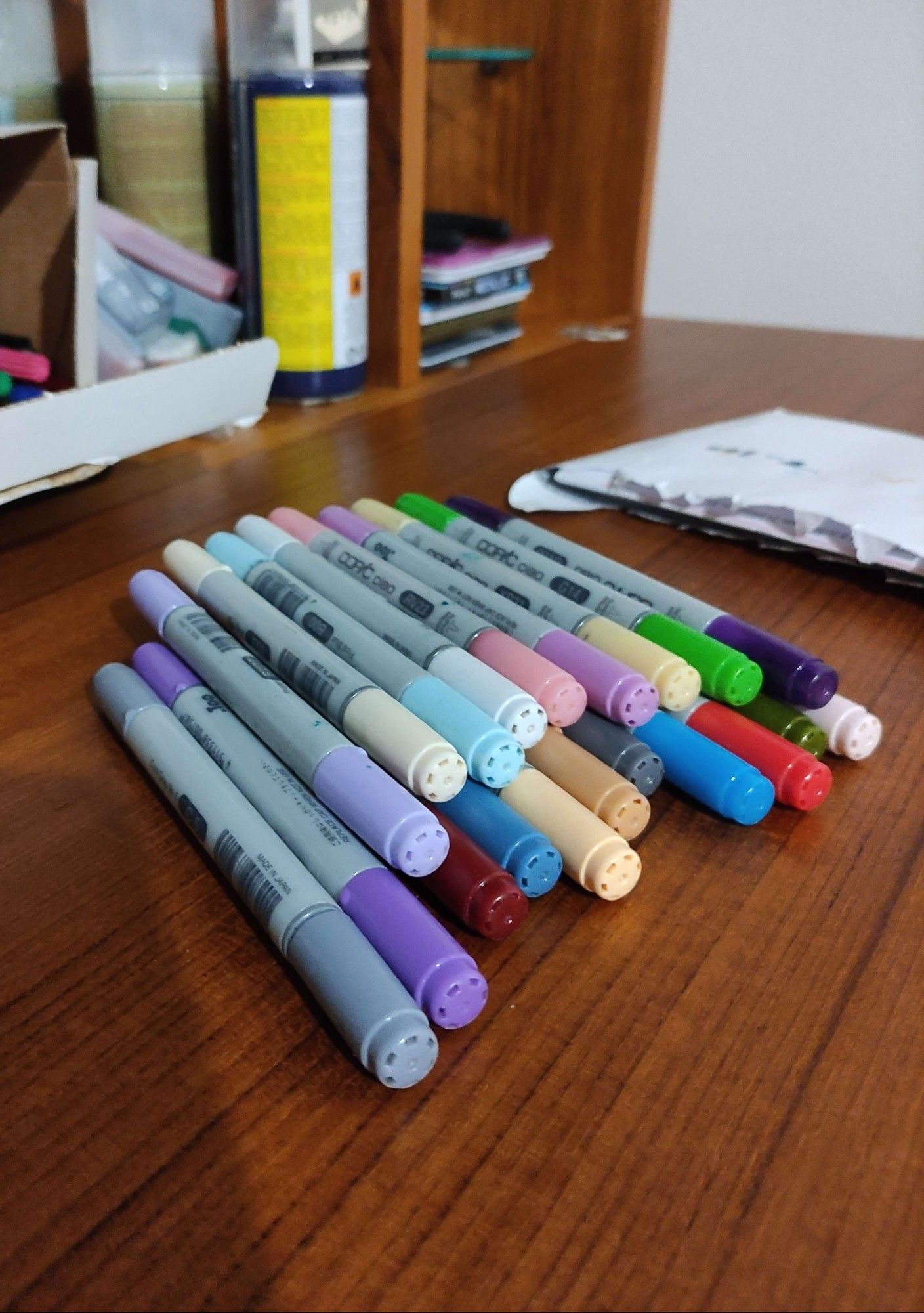 A photo of a small pile of Copic markers on a dark, wooden bookshelf. I'm counting 20 markers. I believe this is all I ever had. A lot of cool tones because I guess I've always preferred them over warm ones, heh.