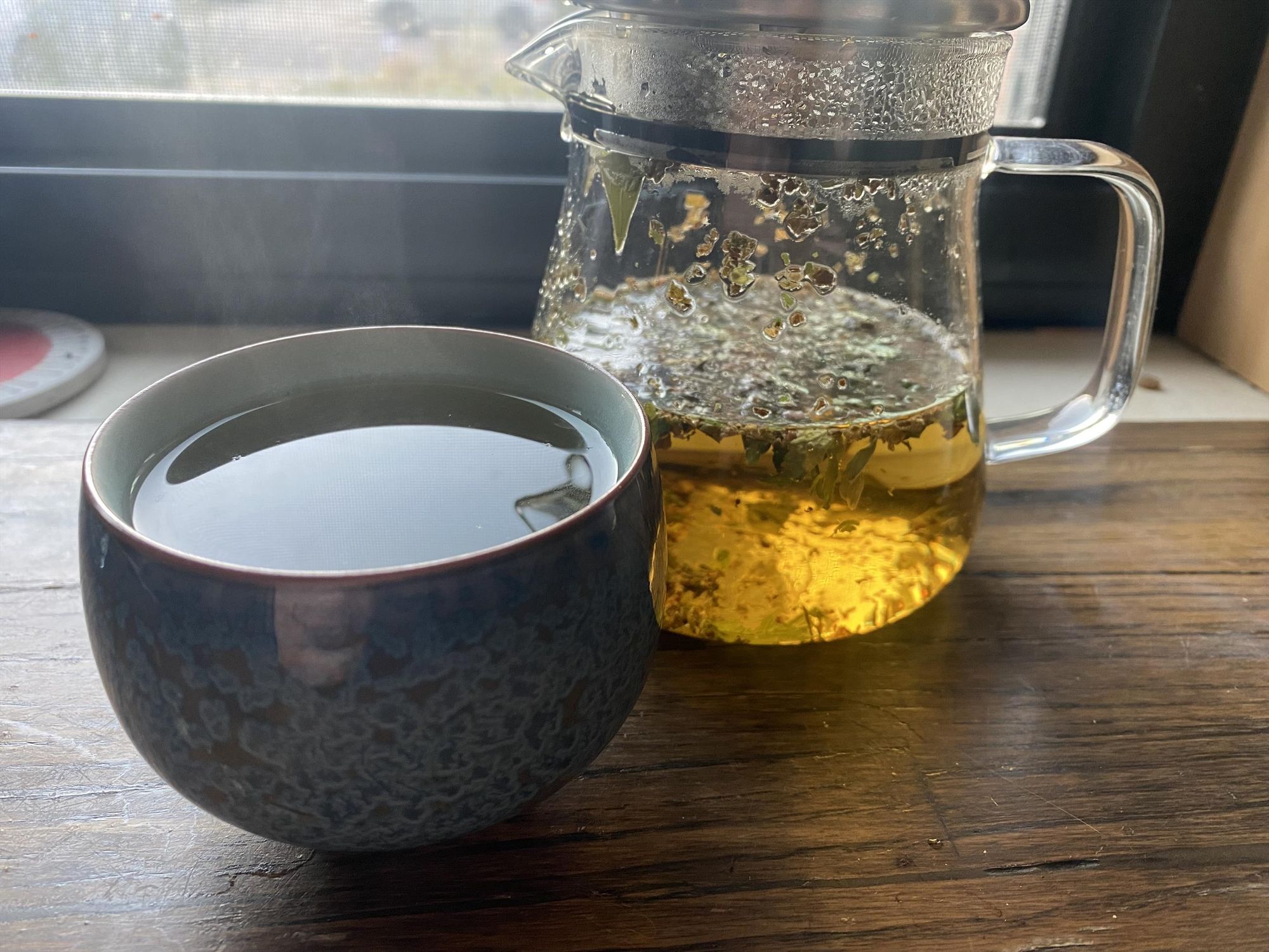 A glass teapot containing broken leaves and a dark golden liquid; a blue teacup with cellular-looking swirls.