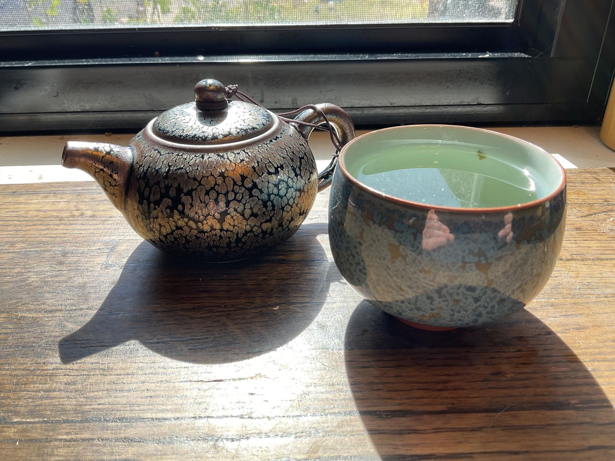 A blue swirled cup filled with a pale green-gold liquid and a gold tea pot, both gleaming in the sun.