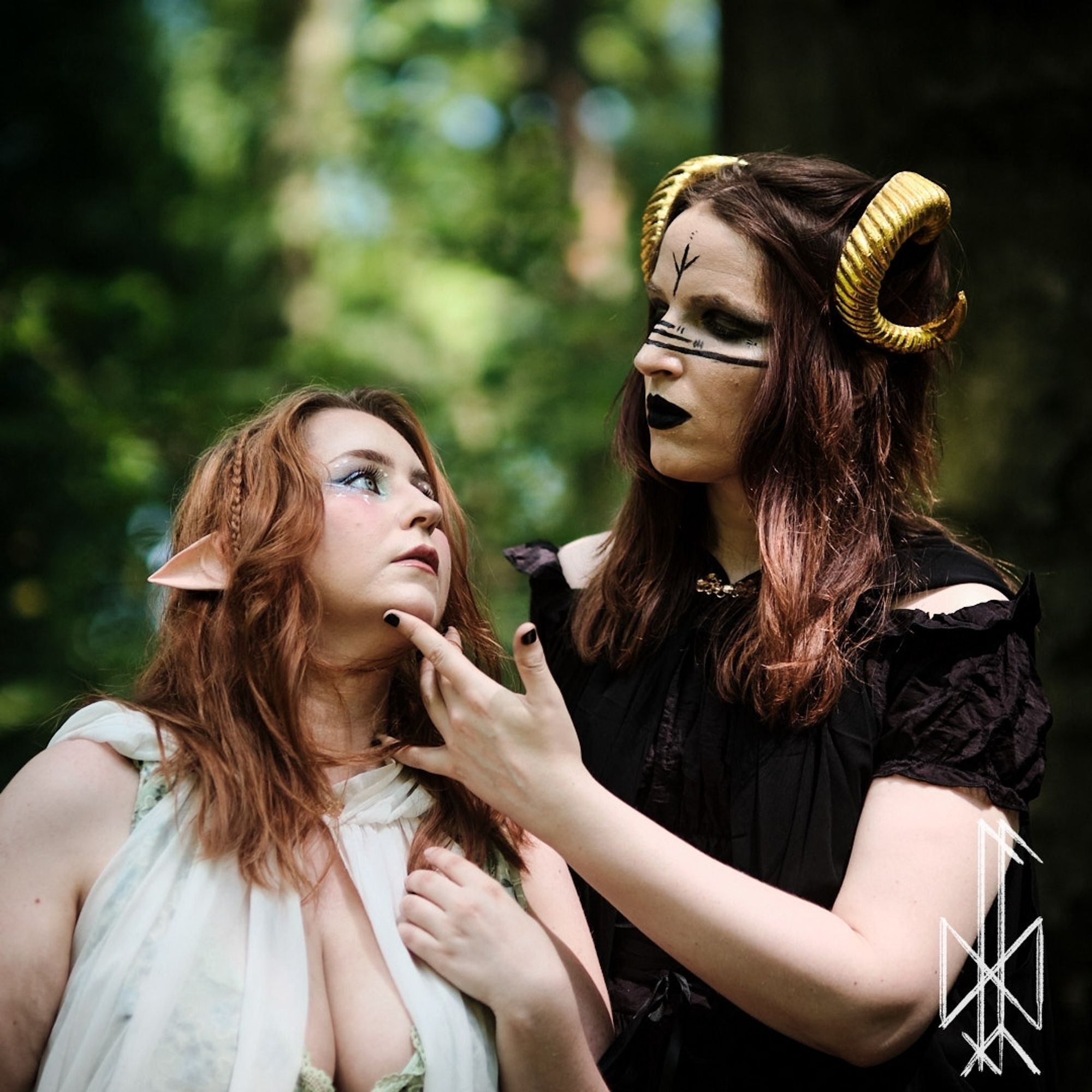 Picture of Lili and her wife Crystallora in the woods. Lora is dressed in a Green Dress and White Cape, with bright Makeup, braids in her reddish Hair and Elven Ears. Lili is dressed in a clad black Dress with cold Shoulders, a black Cape, and her makeup is a White/black Pagan Look with a Rune on her forehead. Lilis dark Hair is adorned with golden demon Horns. They stand next to each other, halfway turned to another and Lili is Lifting Loras Chin gently with a demanding Expression.