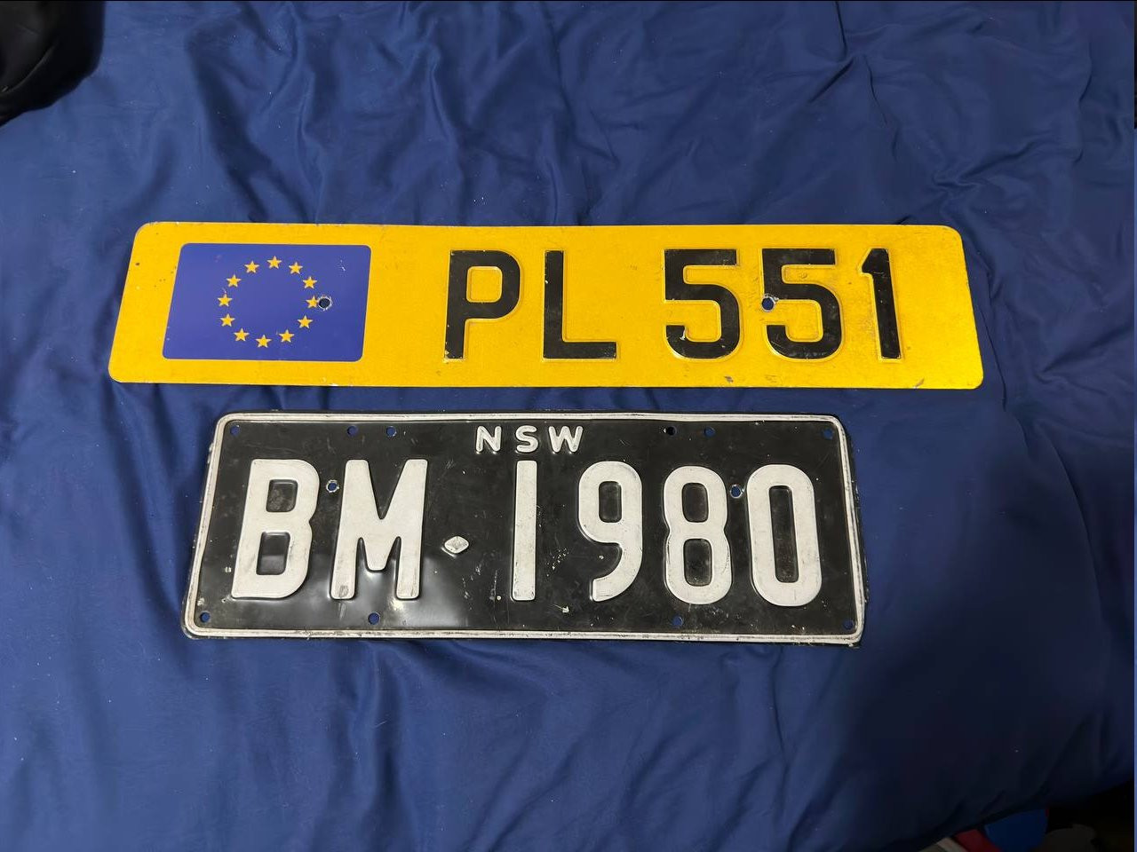 Two license plates: on top, a yellow Luxembourg plate from 1988 with the Flag of Europe on the left and the number PL 551. Below, a black plate with white characters from New South Wales, Australia issued between 1937 and 1951, the number is BM-1980.