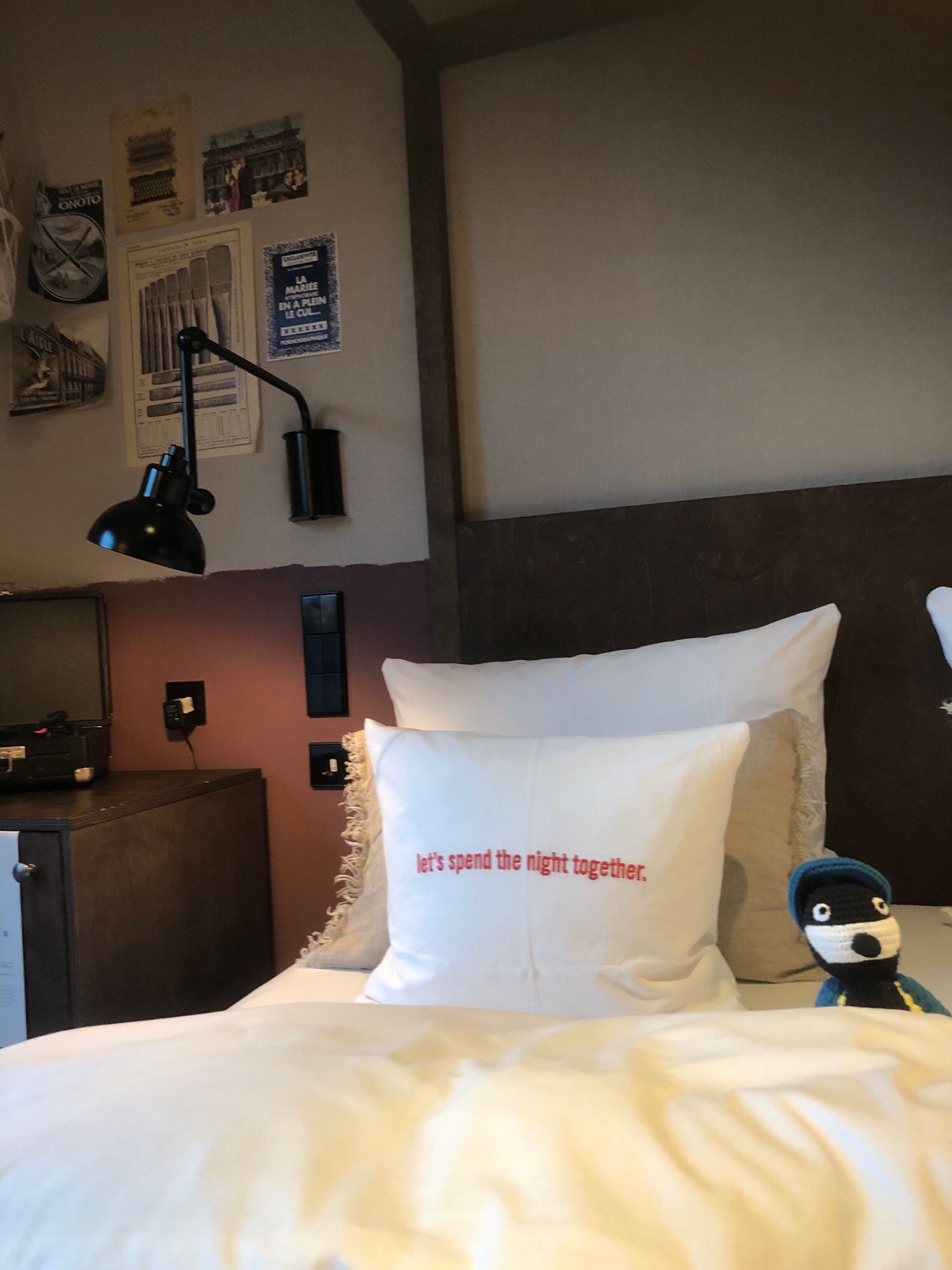 Vorderes Ende meines Hotelbettes mit mehreren Kopfkissen. Auf dem vordersten steht „let’s spend the night together“. Rechts sitzt eine kleine gestrickte Ente mit Baskenmütze. Links ist ein Stück der Minibar und eine Lampe zu sehen.