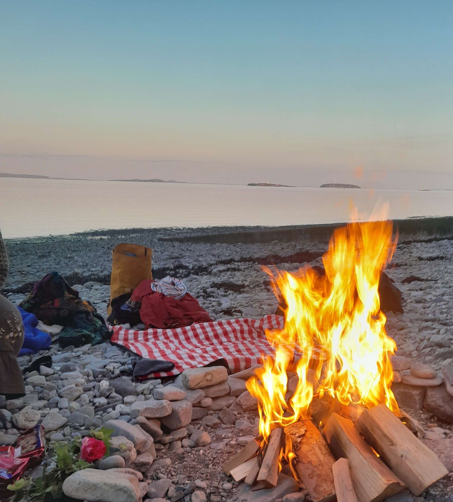 of birch wood and kindling I can't identify, burning in a ring of stones on a pebbly beach. strewn around are flowers, picnic blankets, bags and so on. the sea and sky are lit with pastel dusk colours (blue, orange and pink) as the sun goes down behind frame. across the waters there are several islands