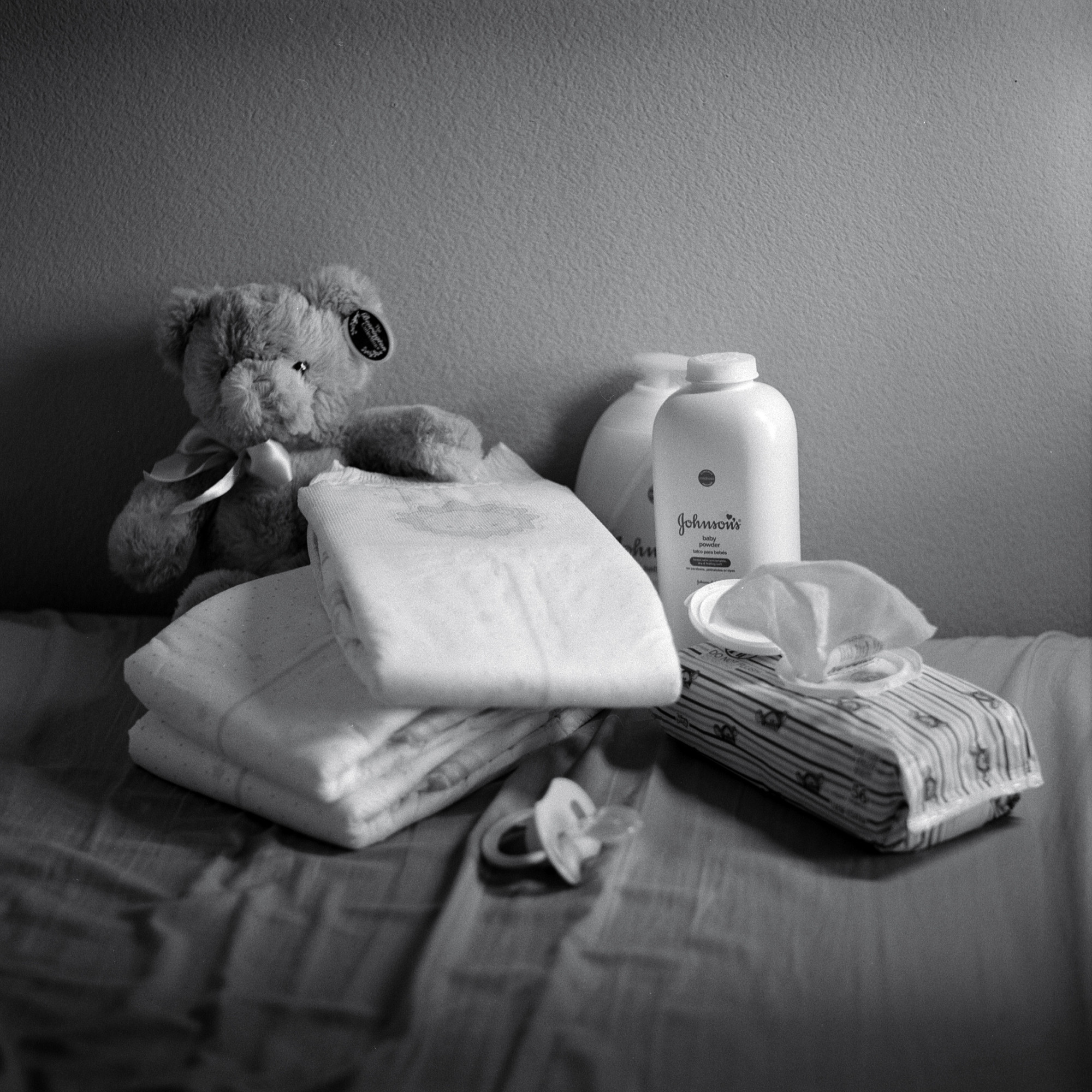 Black and white image of an assortment of little things. Diapers, a teddy bear, wet wipes, a pacifier, babypowder and baby lotion. 