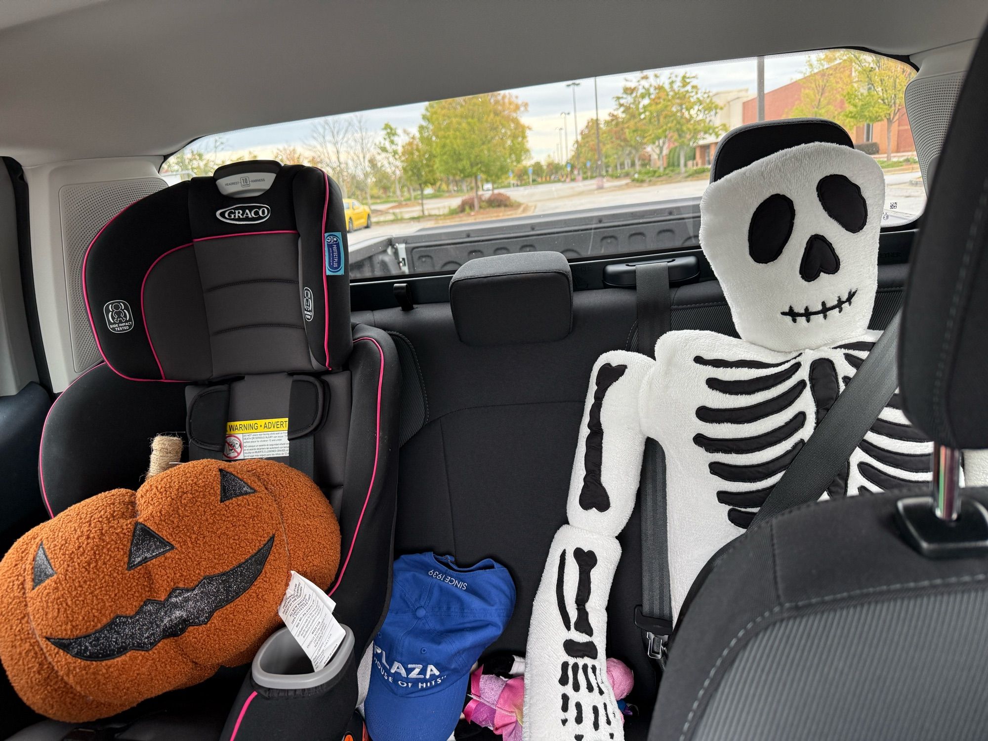 A plush jackolantern in a car seat beside a seat-belted 5ft skeleton pillow.