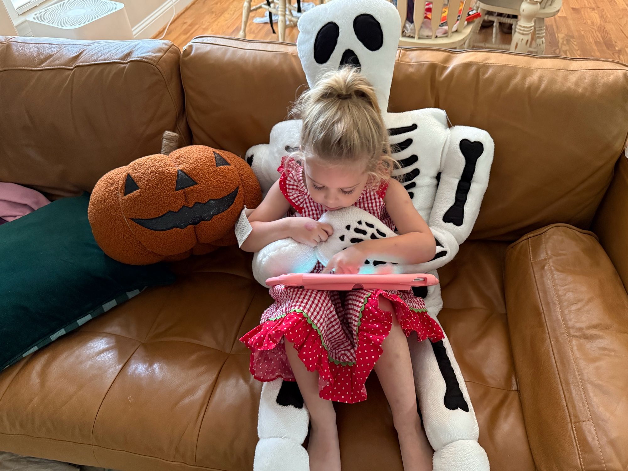 My toddler sitting in the lap of a 5ft plush skeleton pillow beside a plush jack-o’-lantern