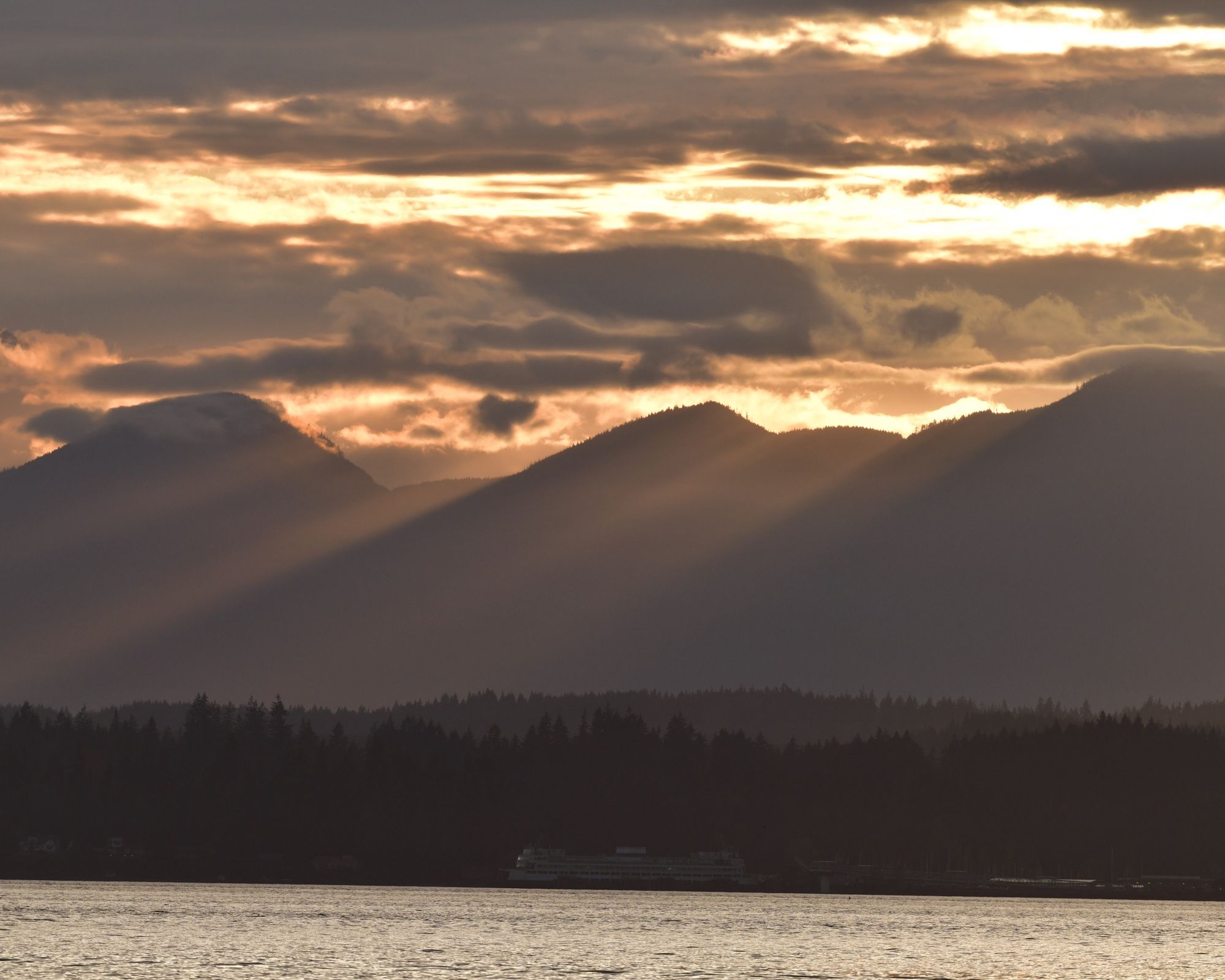 The Olomouc Mountains in golden sunset lit, with beams of sunlight shining through the passes. Above, golden clouds. Below, Puget Sound and a row of dark trees in front of the mountains.