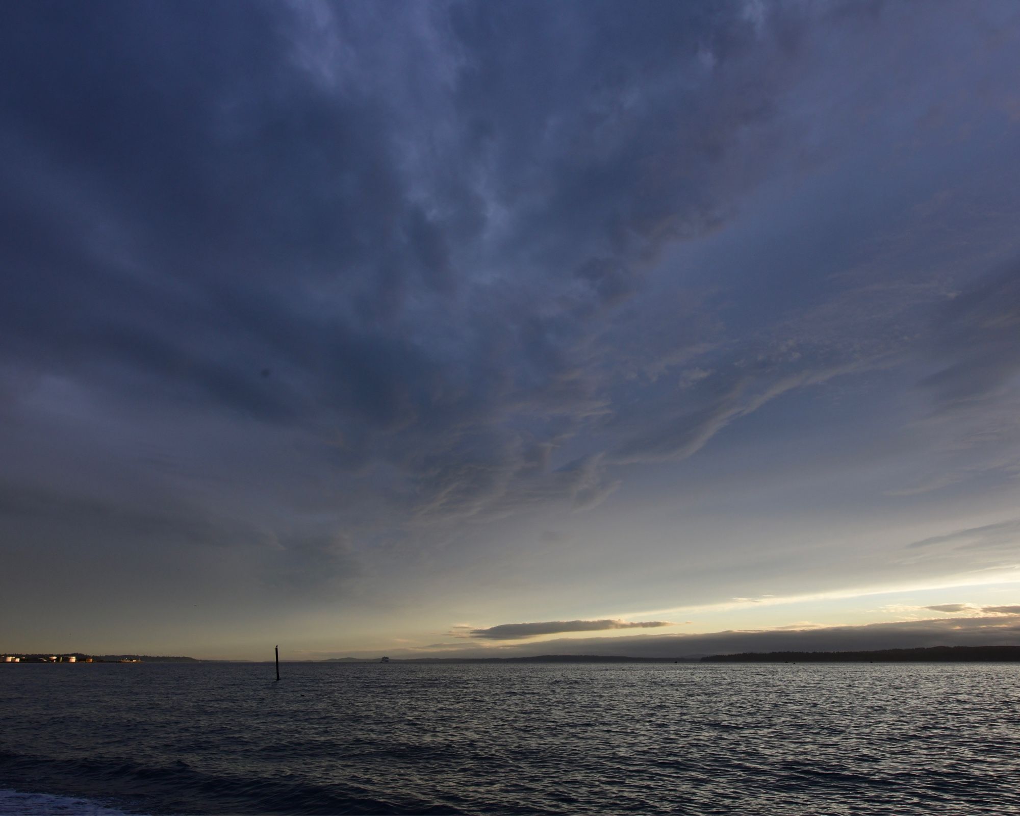 Puget Sound, dark below a cloudy sky. The left is in dusk, dark an almost foreboding. The clouds are dark and seem to be writhing. To the right, sunset and a wedge of golden sky slices in between water and clouds, tinting everything.