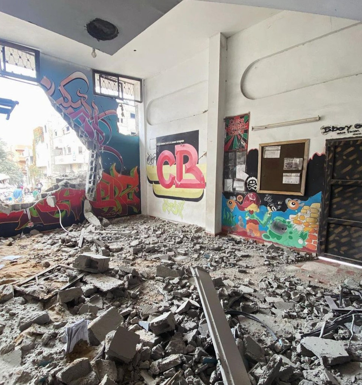 Destroyed Camps Breakerz space, the foreground littered with broken pieces of cement, a huge hole through a wall painted with a mural shows more destruction outside the building. The in-tact wall is also painted colorfully with cartoon characters and the letters 